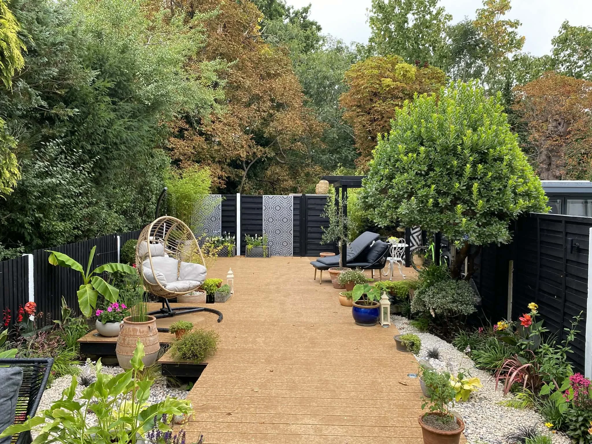 A sleek modern back garden with decking and plants, outdoor furniture, and a black fence, surrounded by lush trees and plants with varying shades of green