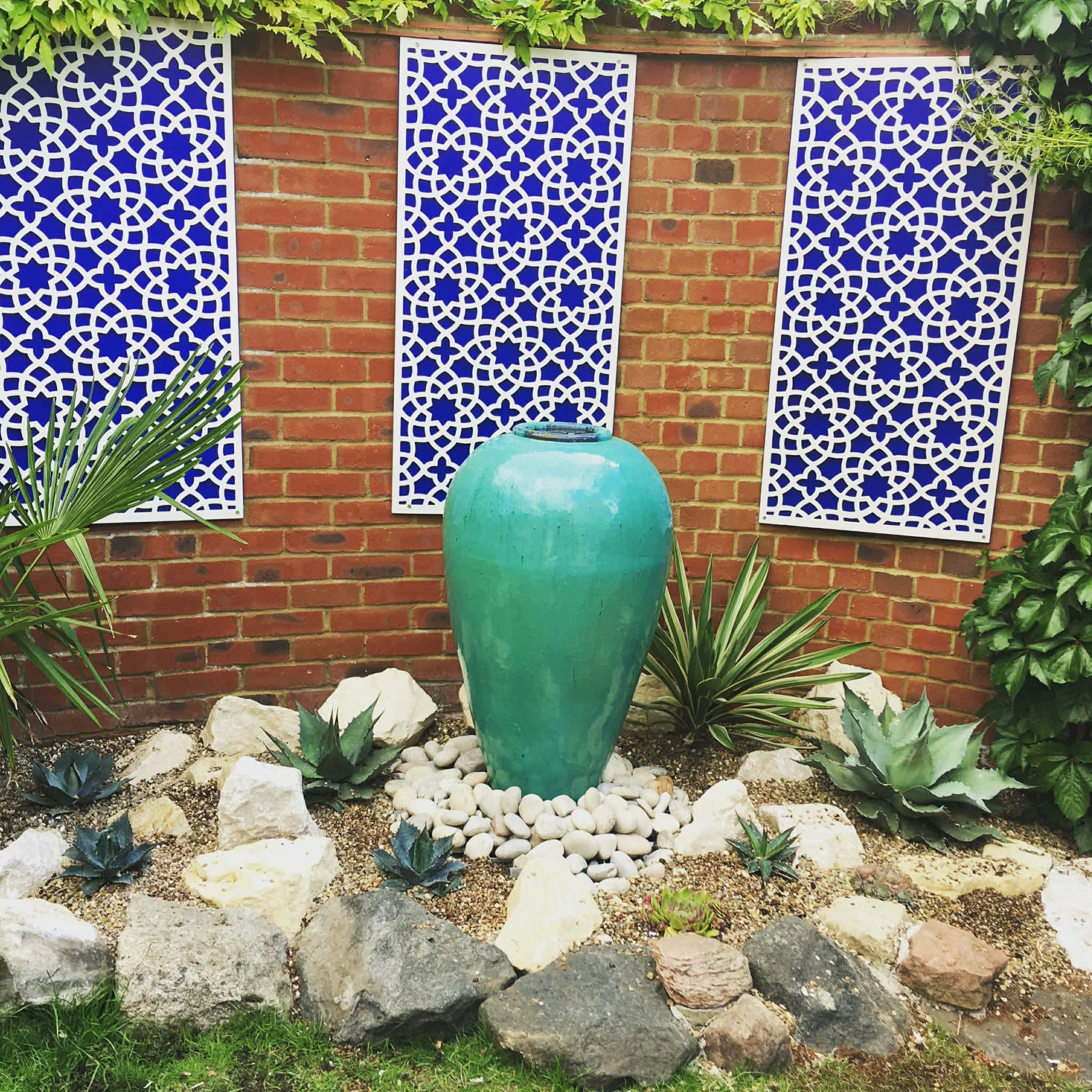 A decorative Moroccan style garden scene with white carved panels on a red brick wall, a large green ceramic pot, and various plants, rocks, and white pebbles in the foreground.