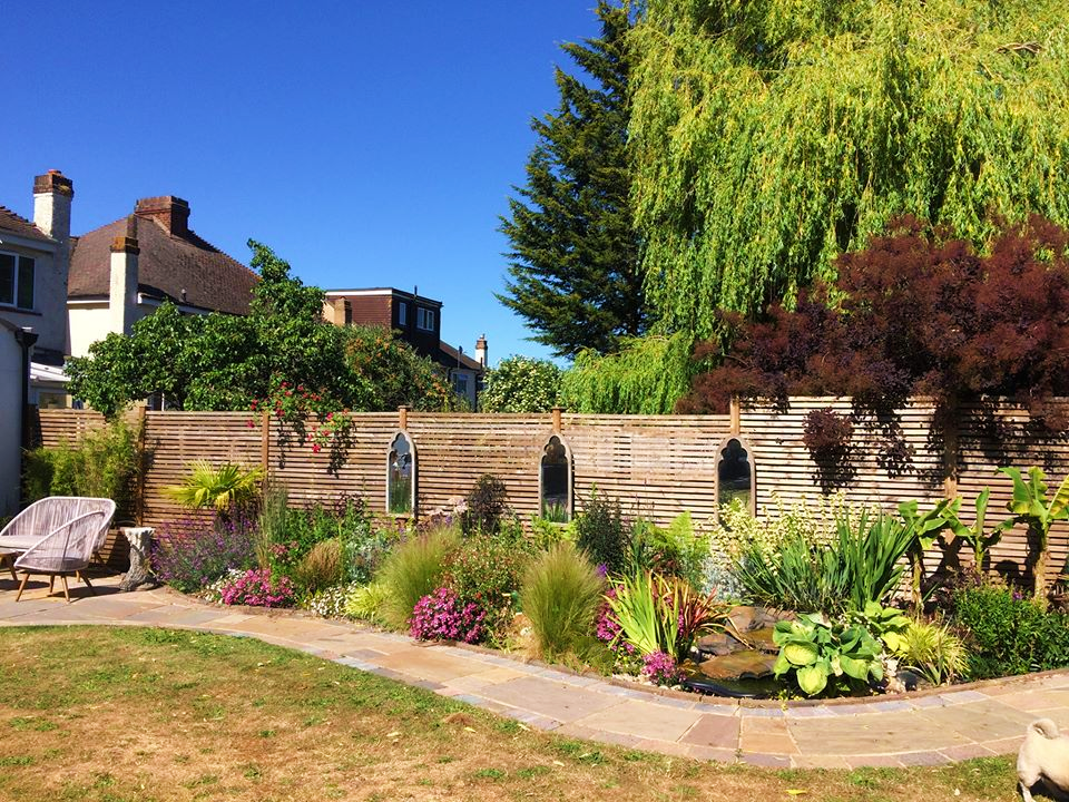 A colorful garden with a variety of flowers and plants, a stone pathway, a wooden fence, and neighboring houses in the background under a bright blue sky.