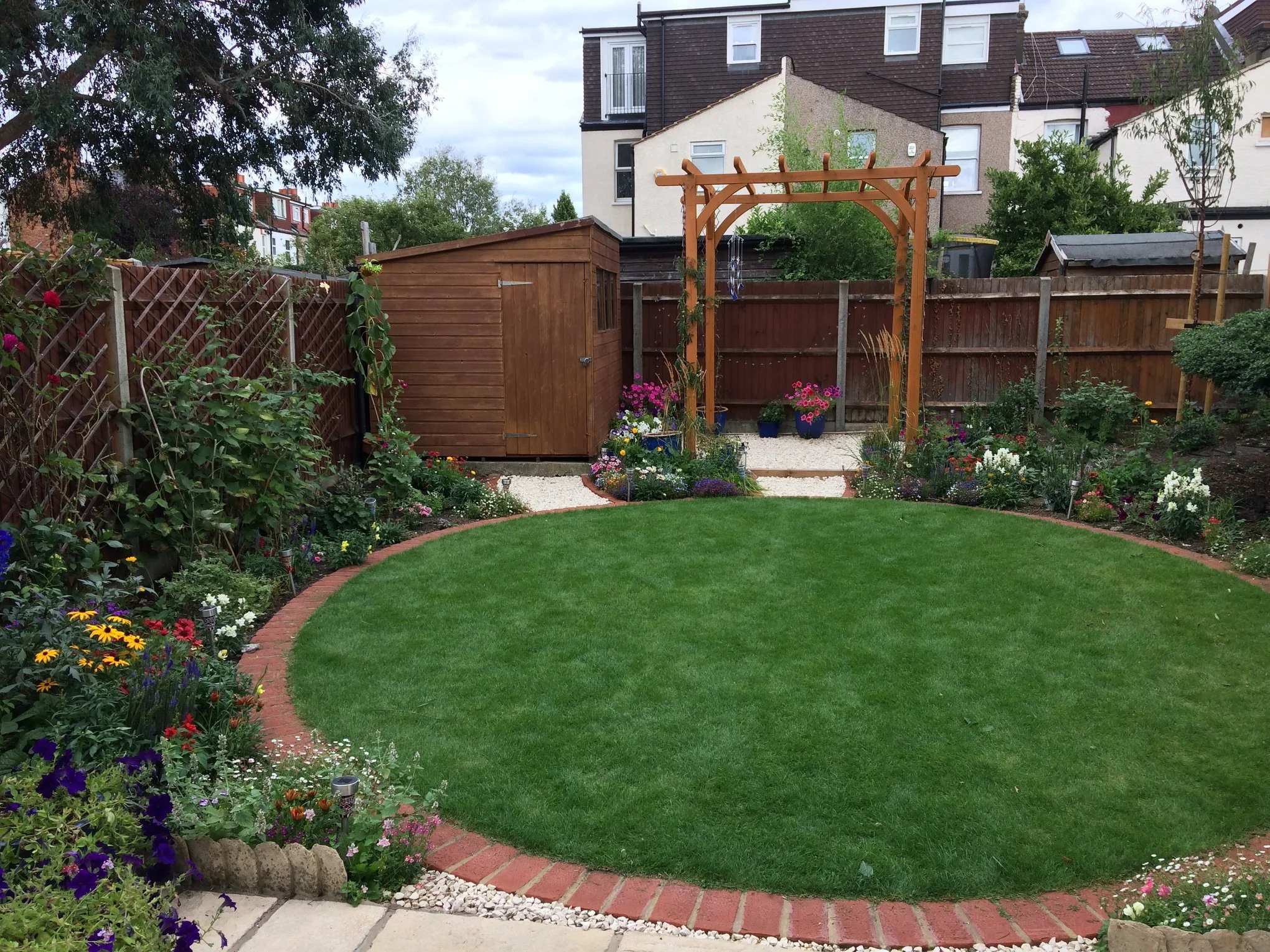 A backyard garden with a well-maintained green lawn, surrounded by colorful flowers and plants. There is a wooden shed on the left, a wooden archway with hanging decorations in the center, and an assortment of potted flowers nearby. A gravel pathway leads through the garden, and houses with residential windows are visible in the background under a cloudy sky.