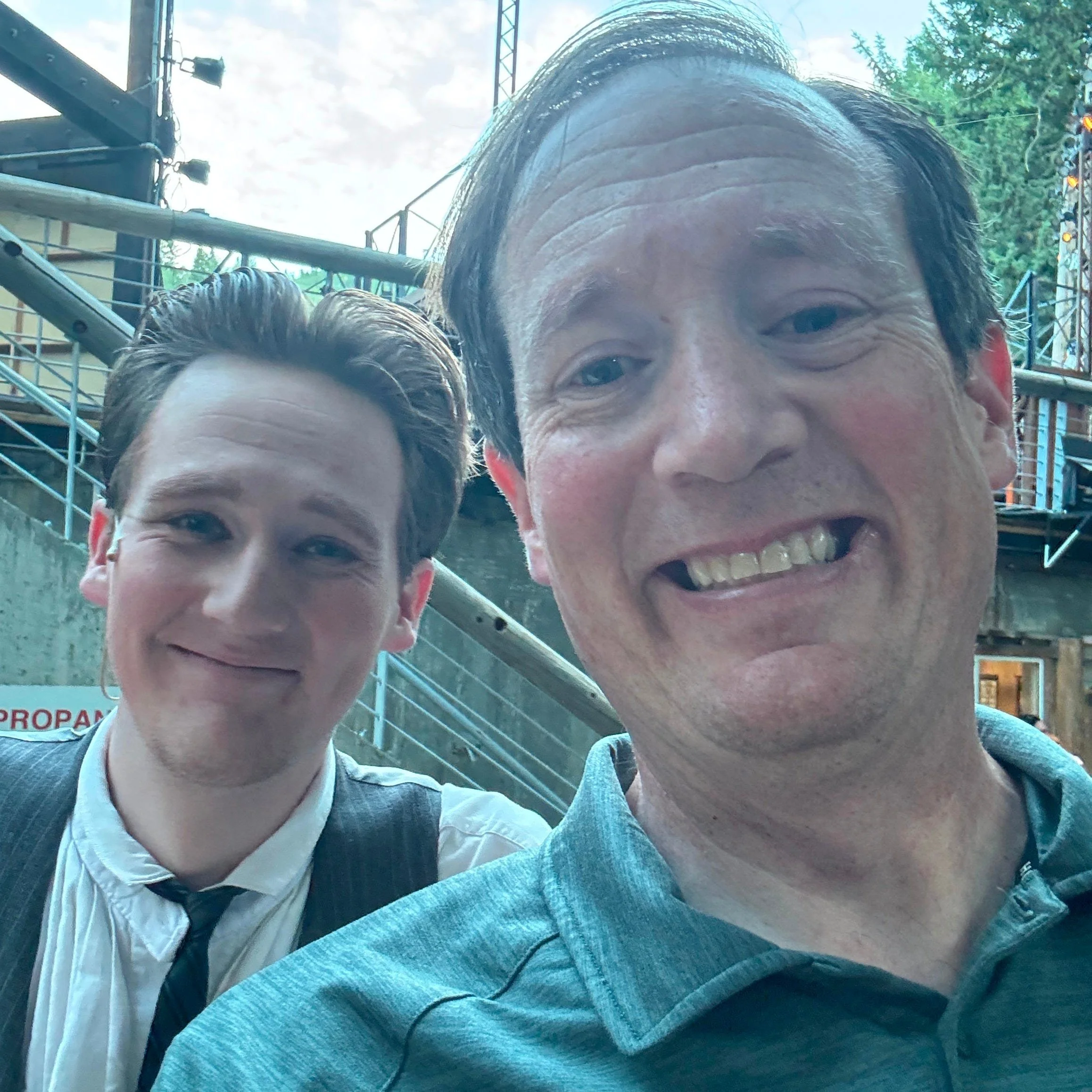 Two men smiling and taking a selfie outdoors, with trees and metal fencing in the background.