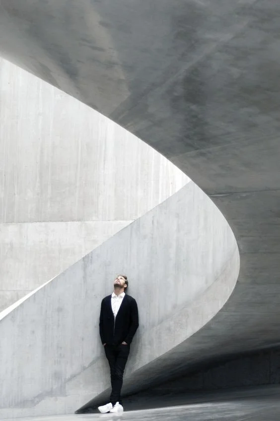 Man in black jacket, pants, and white shoes leaning against a curved concrete architectural structure with his head tilted upward.