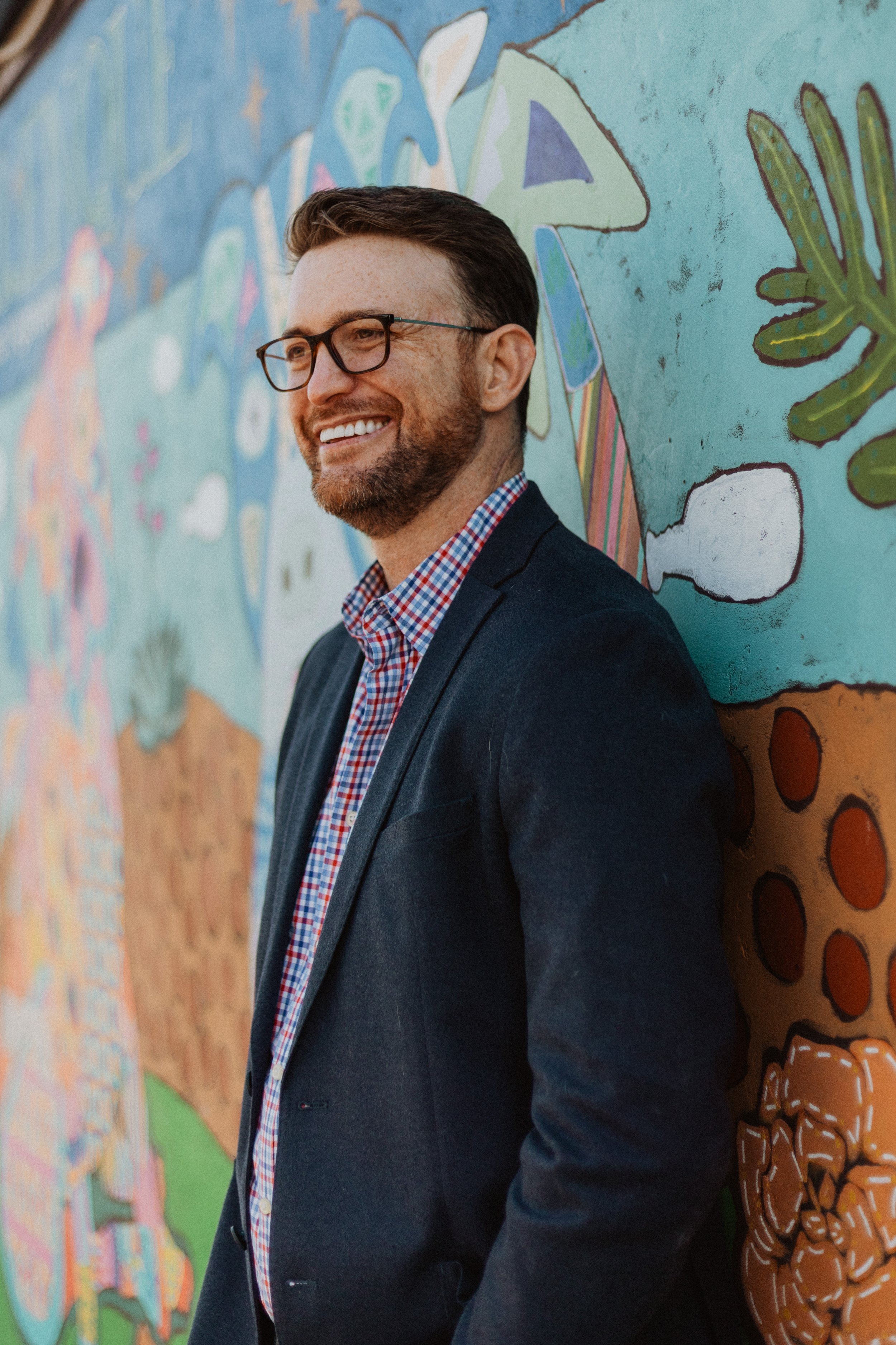 A man with glasses and a beard, smiling and wearing a checkered shirt and blazer, leaning against a colorful mural with abstract shapes and patterns.