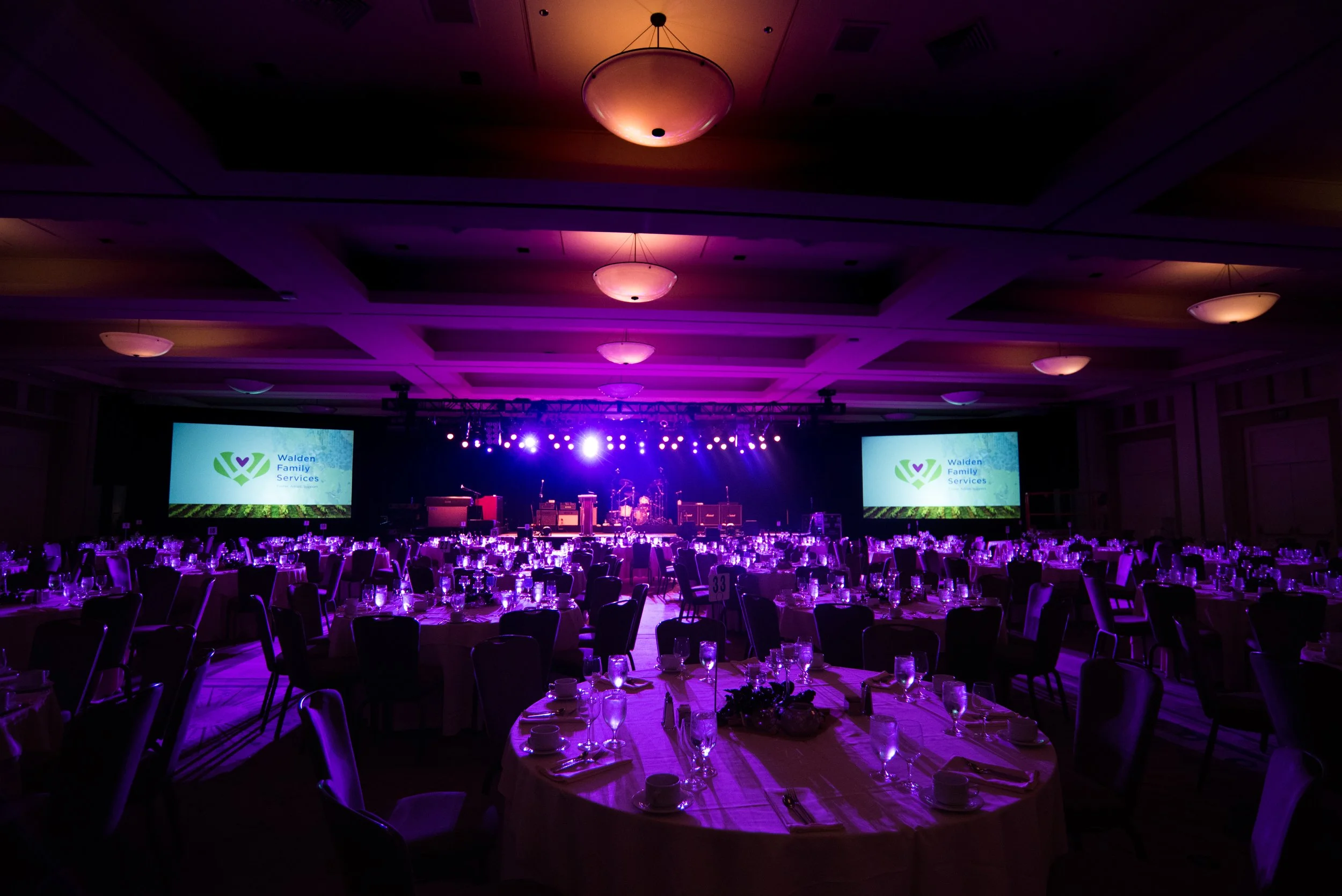 A large banquet hall set up for an event with round tables covered with white tablecloths, glasses, and plates, illuminated with purple lighting. There are two large screens displaying 'Walden Family Services' logo on either side of a stage with musical instruments and bright stage lights.