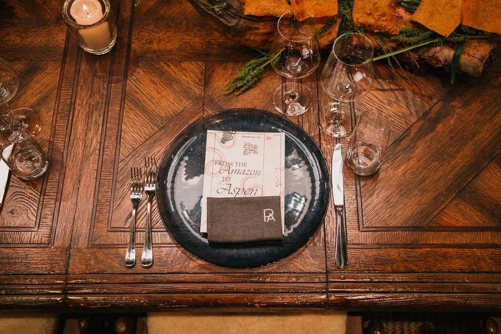 Empty dinner plate with a menu card placed on top, set on a wooden table with silverware, glasses, and a candle.