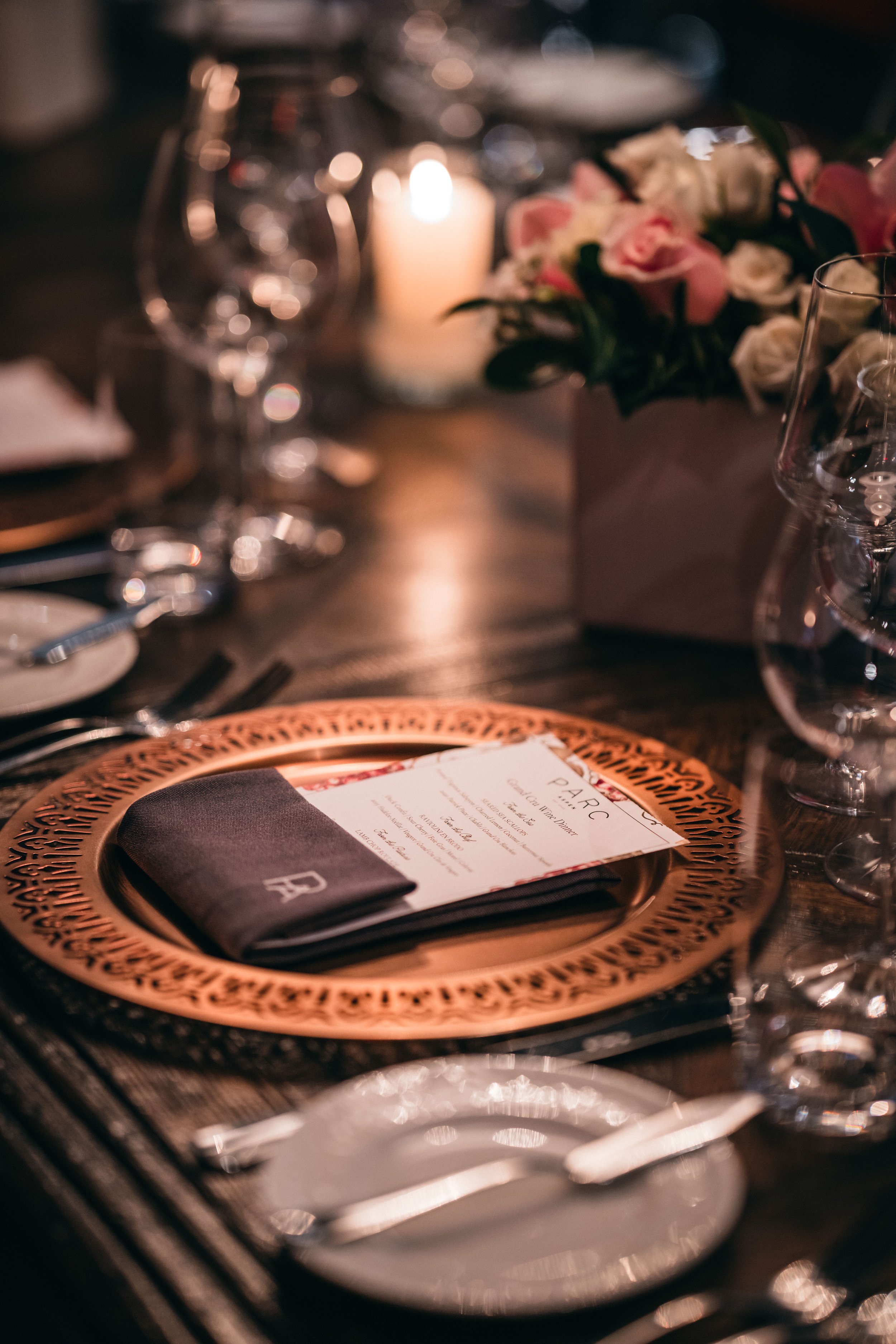 A dinner table setting at PARC Aspen, local fine dining destination, with a decorative charger plate, a folded menu, and a black napkin with a monogram, set for a formal occasion. In the background, a lit candle and a floral centerpiece are visible.