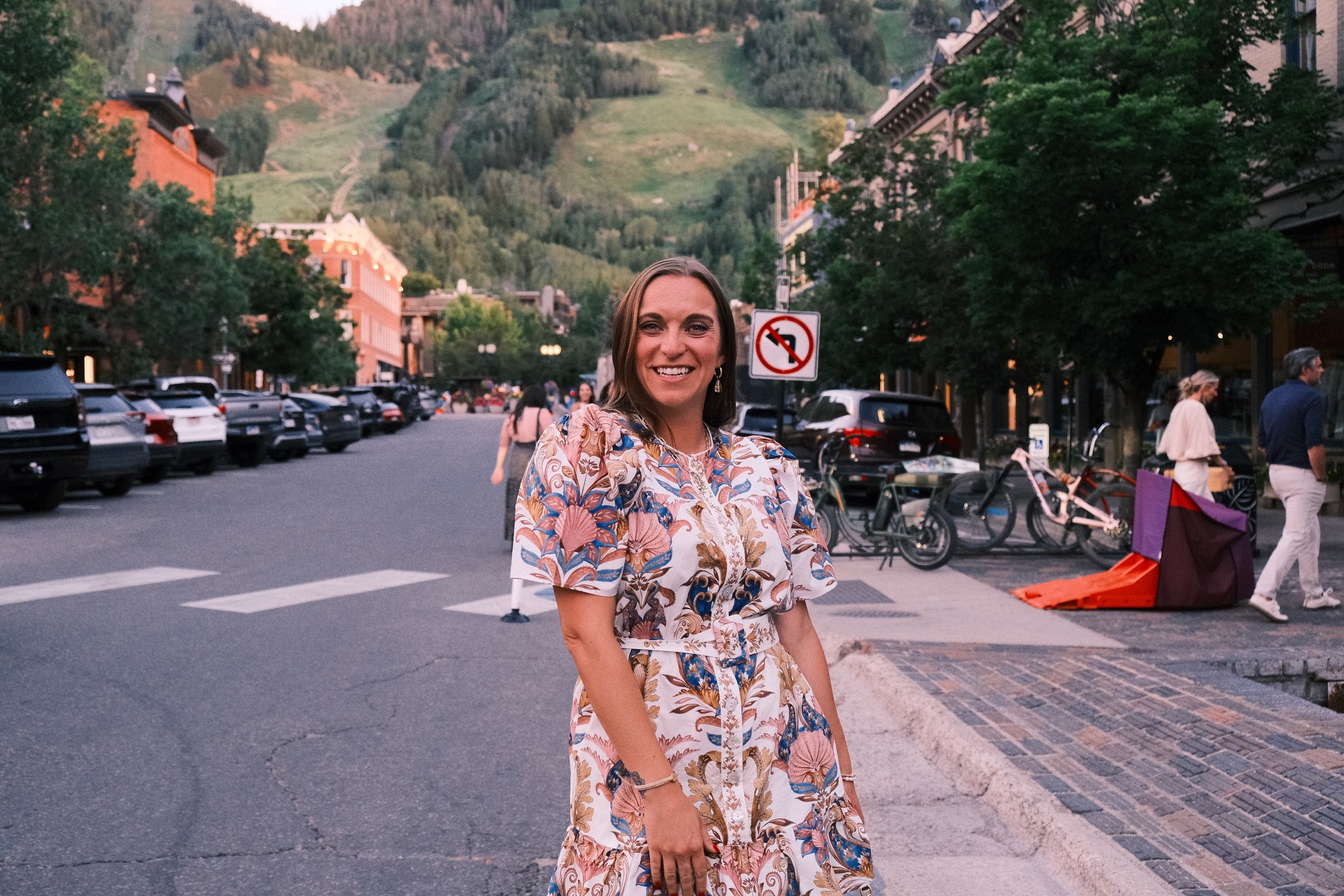 A smiling woman with long brown hair standing on a city street with parked cars, trees, and buildings in the background. There is a mountain with greenery behind her, and a 'no left turn' traffic sign is visible.