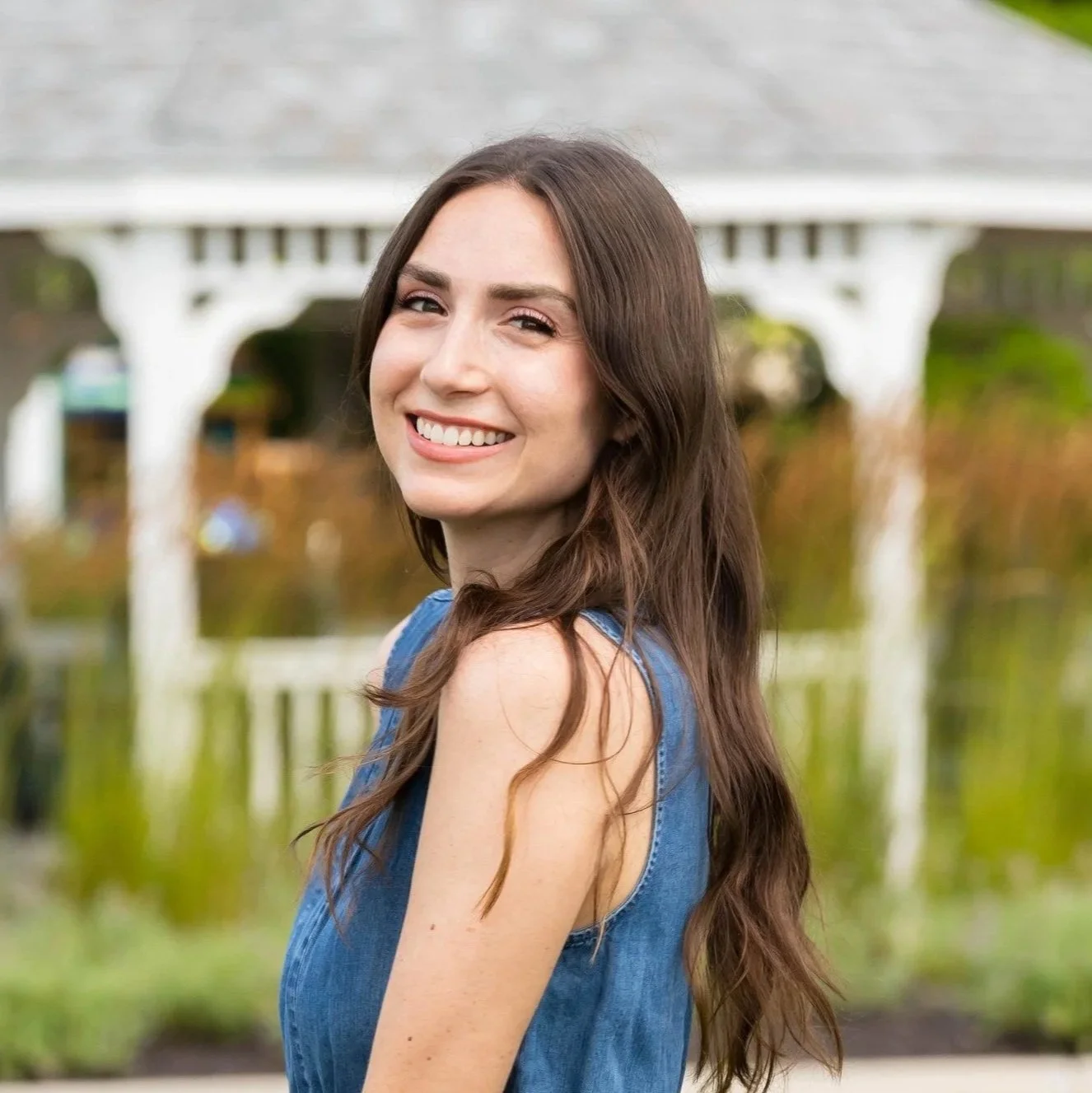 Smiling woman with long brown hair, wearing a sleeveless denim top, standing outdoors near a pond, with a white bridge in the background.