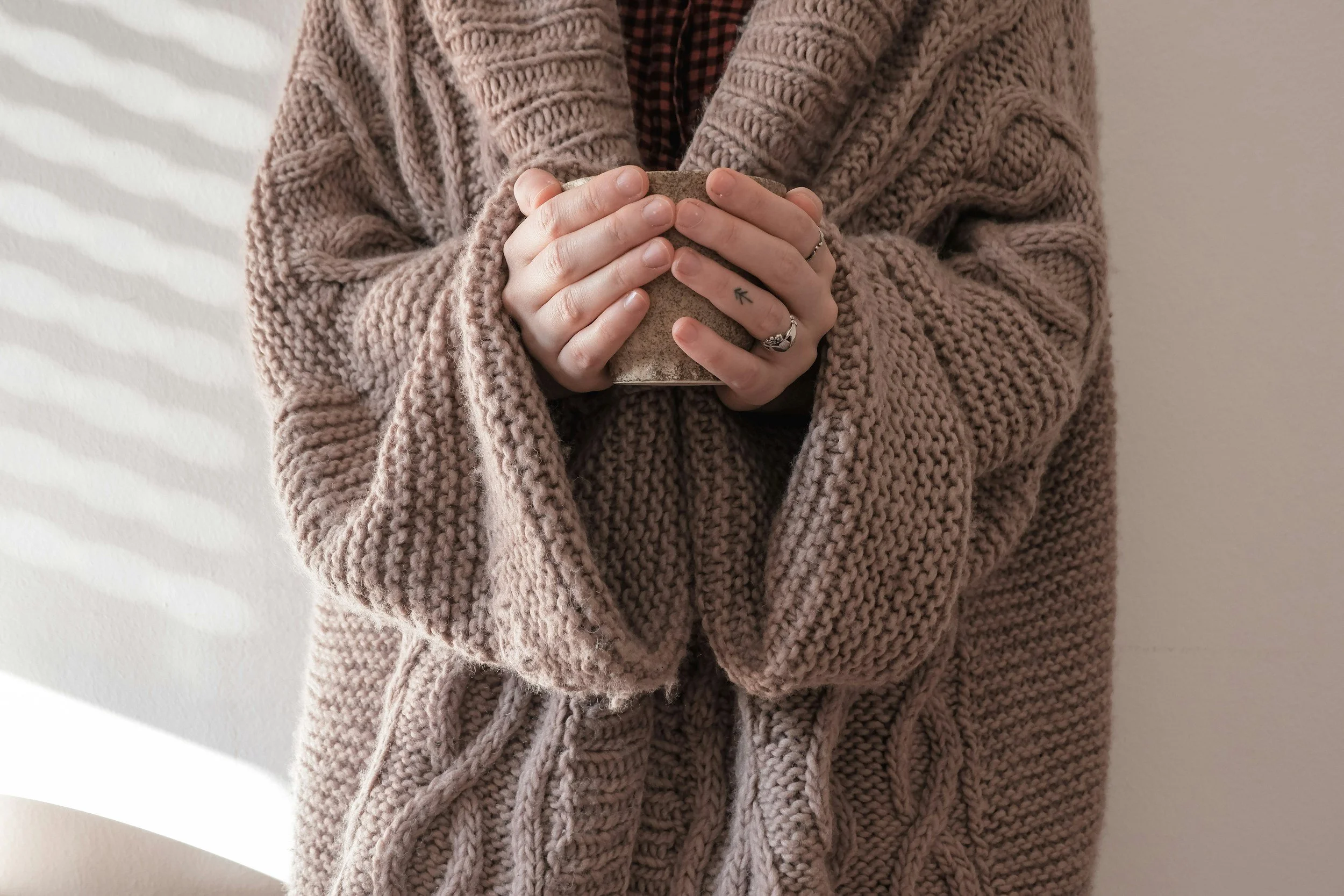 Person wearing a chunky knitted sweater with their hands clasped, holding a small stone or cup. The person has rings and a small tattoo on their fingers. Background is plain and light-colored.