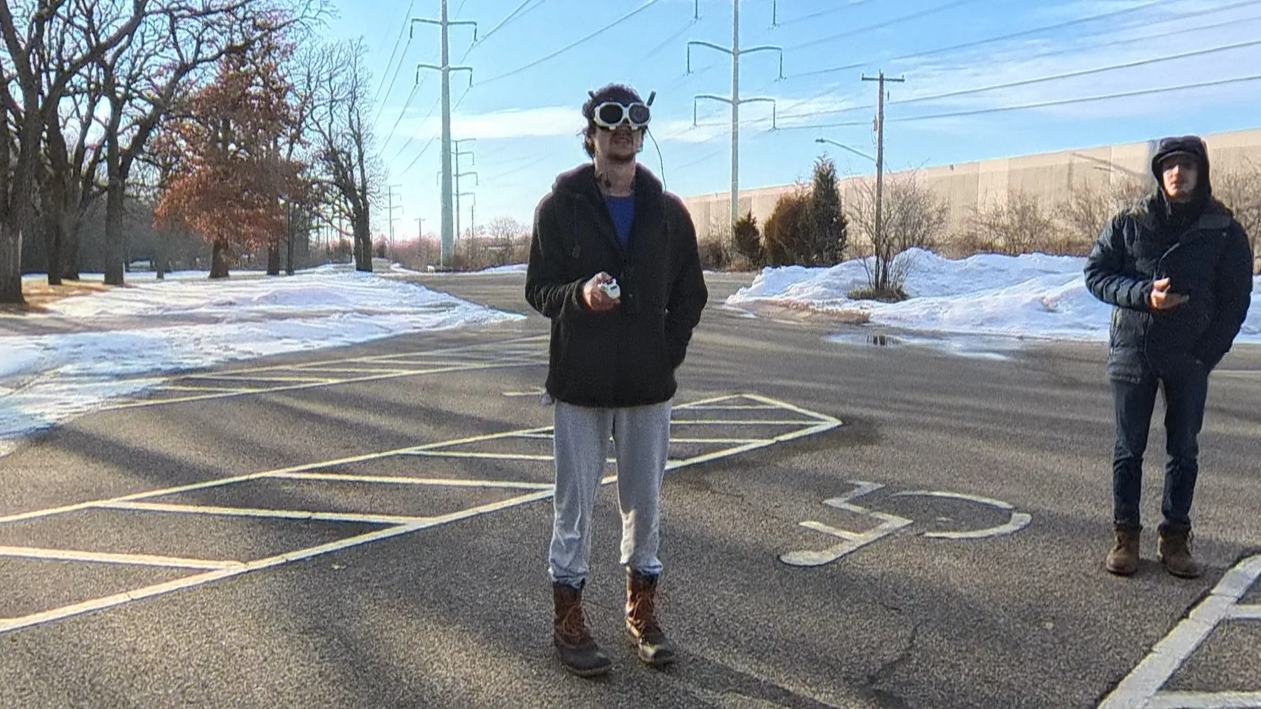 Two young men standing in an empty parking lot in winter, with snow on the ground and bare trees in the background. One man in the foreground wears goggles, a black jacket, gray sweatpants, and boots, holding a small object. The other man in the background wears a black jacket, jeans, and boots, holding a phone.
