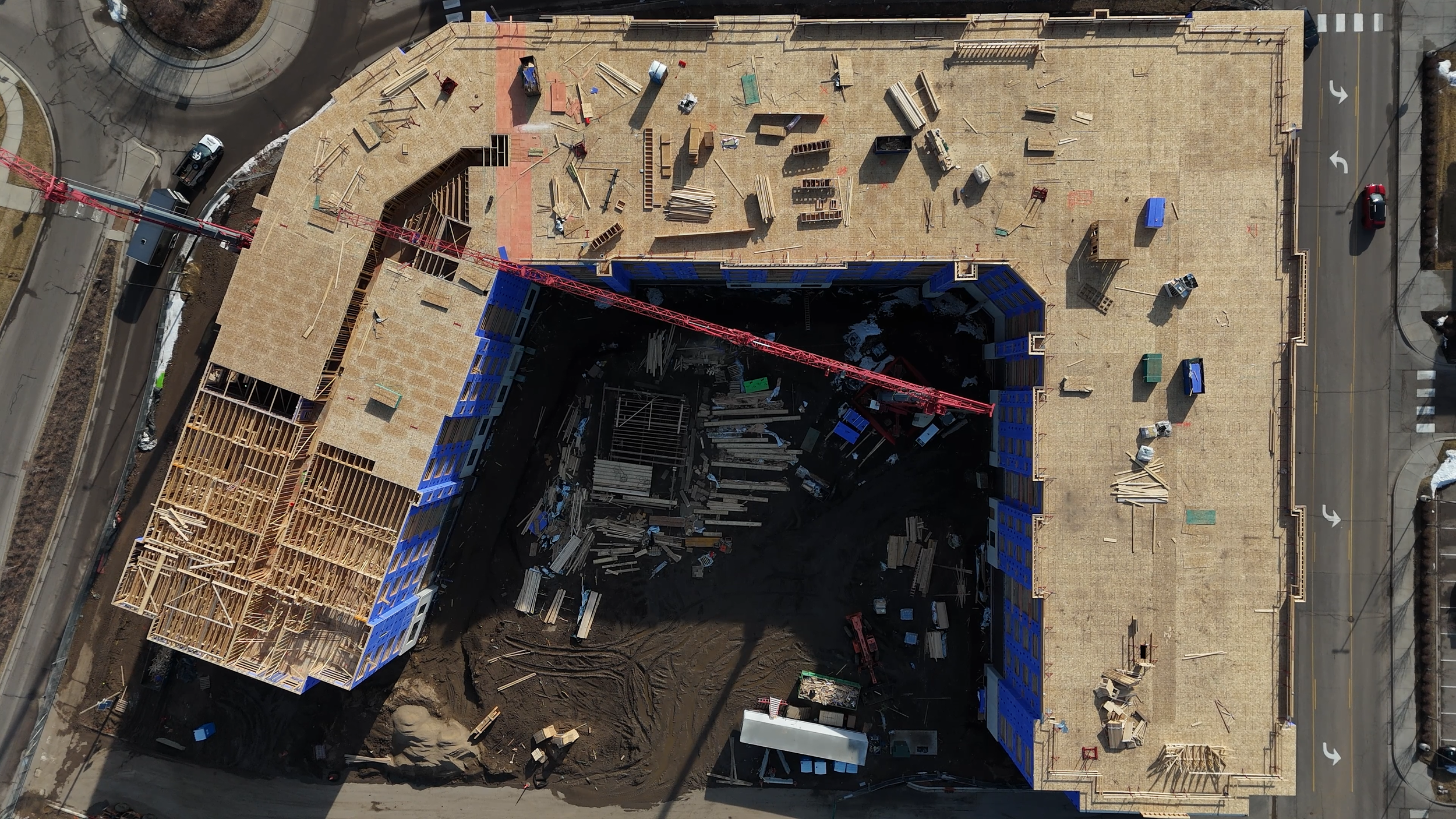 Aerial view of a construction site with a partially built multi-story building. The building's roof framing is visible, and construction materials and equipment are scattered around. A large red crane extends across the site.