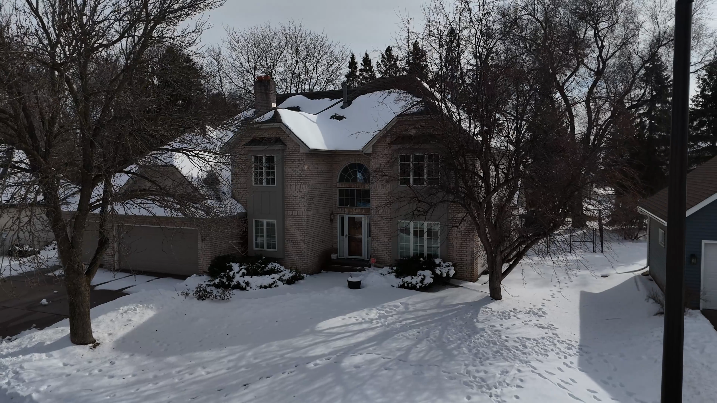 A two-story brick house with a snow-covered front yard, leafless trees, and a driveway on the left side during winter.