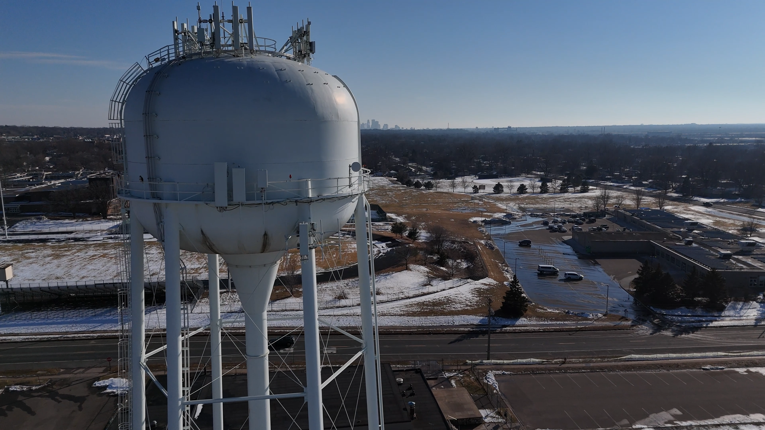 A large white water tower with antennas on top, set against a background of a partly snowy landscape and a clear blue sky.