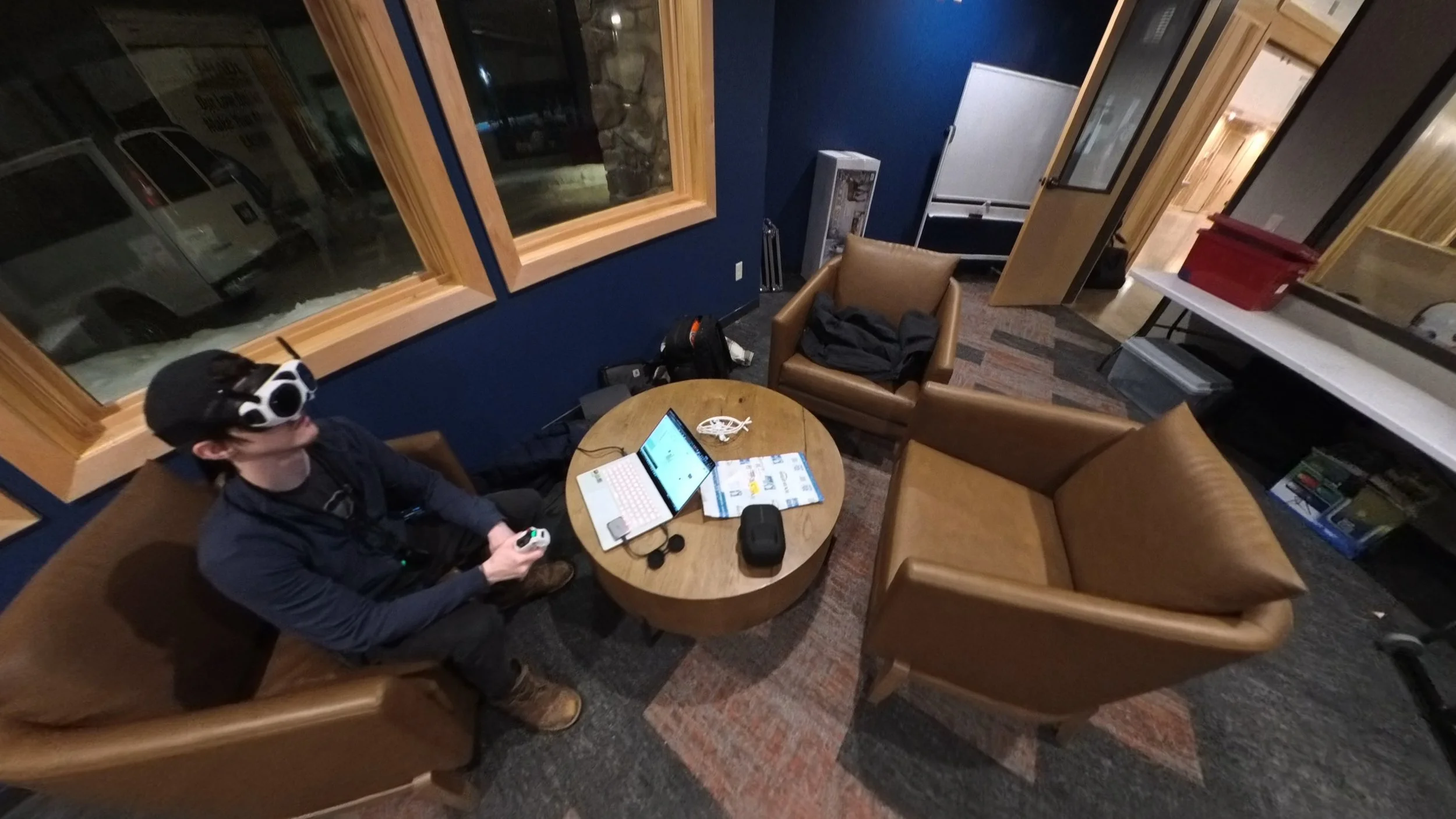 A young man with glasses and a hat wearing virtual reality goggles sitting on a brown leather armchair in a cozy room. There is a round wooden table in front of him with a laptop, a magazine, a small black case, and a necklace. The room has dark blue walls, large windows, and wooden accents.