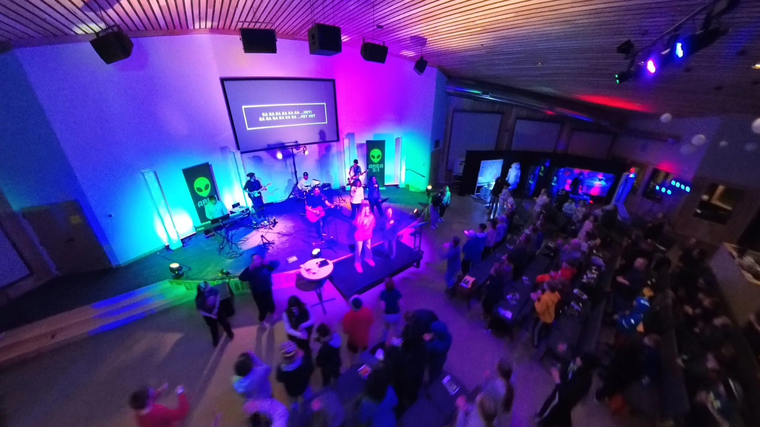 An indoor event with a stage where a band is performing, illuminated by colorful lighting. The audience is gathered in front of the stage, some standing and others seated.