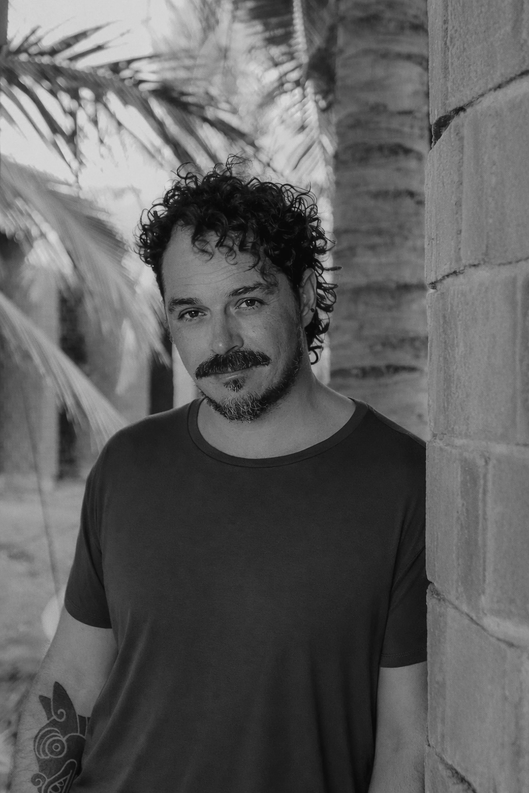 A photo of Ozzy Lusth; man with curly hair smiles slightly while leaning against a brick wall outdoors, with palm trees in the background.