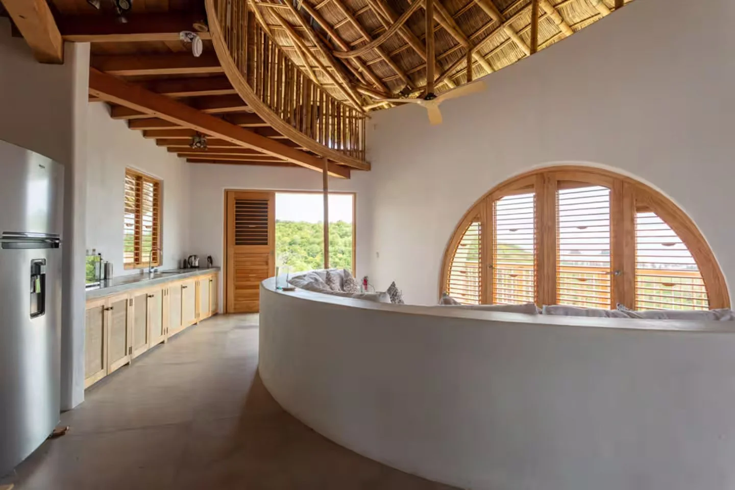 Villa Tortuga; Open-concept kitchen and living area with a curved white wall, large arched window with wooden shutters, wooden ceiling with thatch roofing, wooden cabinets, and a sliding door leading outside to green landscape.