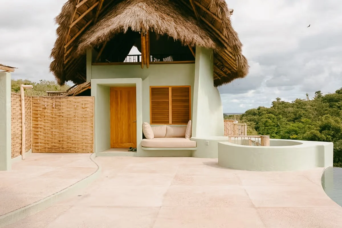 Villa Tortuga; A tropical-style house with a thatched roof, light green walls, wooden door, a window with wooden shutters, a built-in beige cushioned bench, and a circular small wall, surrounded by lush greenery and cloudy sky.