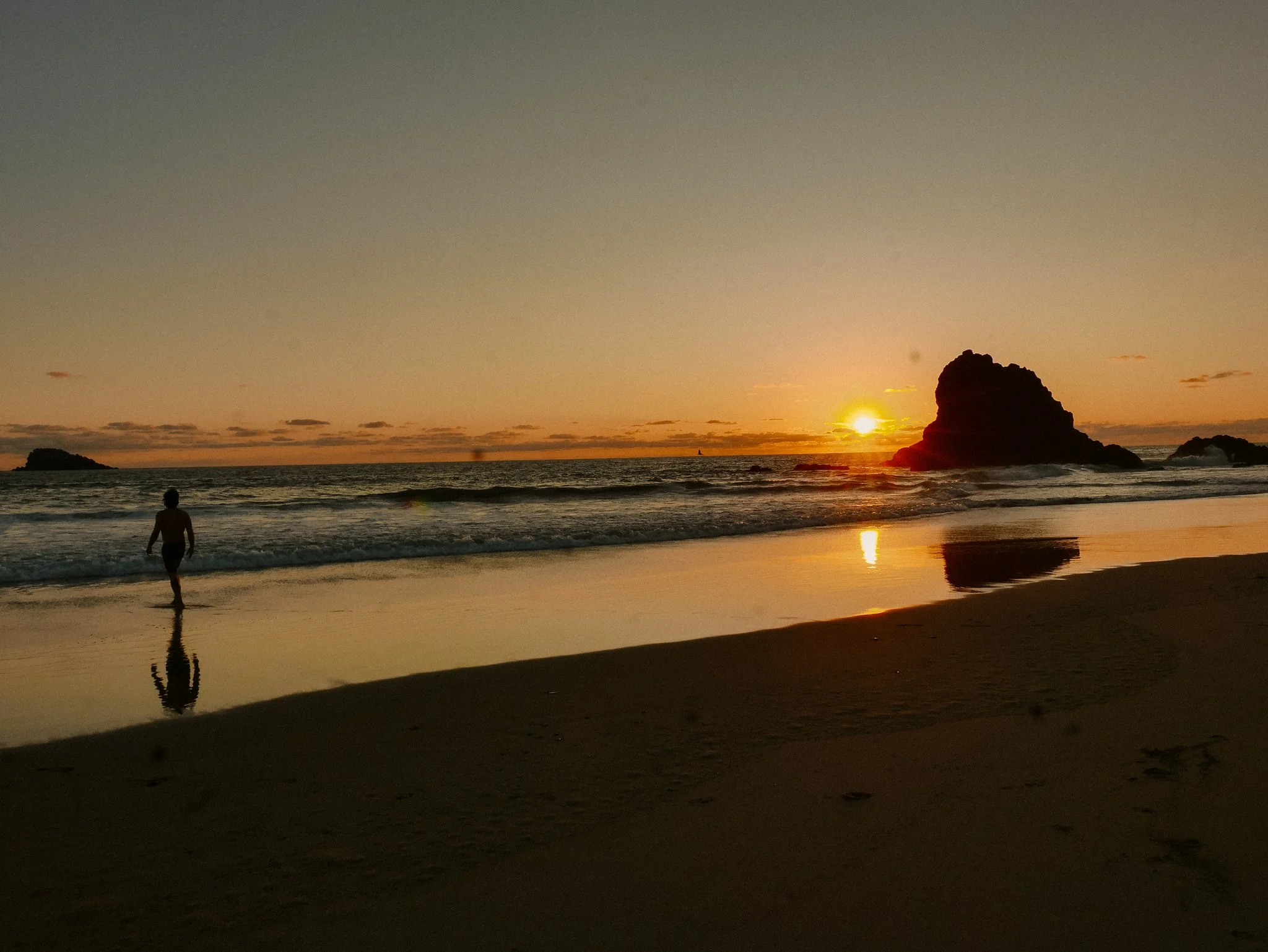Person walking along the shoreline of Arroyo Seco at sunset with large rock formations in the ocean, reflections on wet sand.