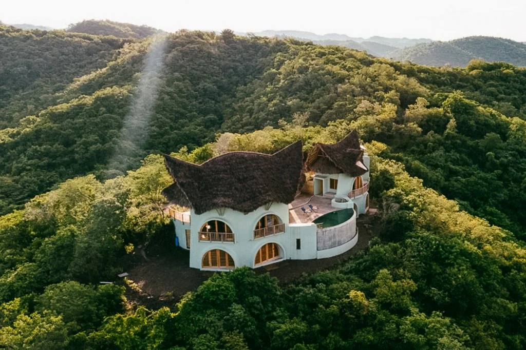 Villa Tortuga; A unique house with a curved white exterior and thatched roofs, situated on a lush green hilltop surrounded by dense forest and mountains.