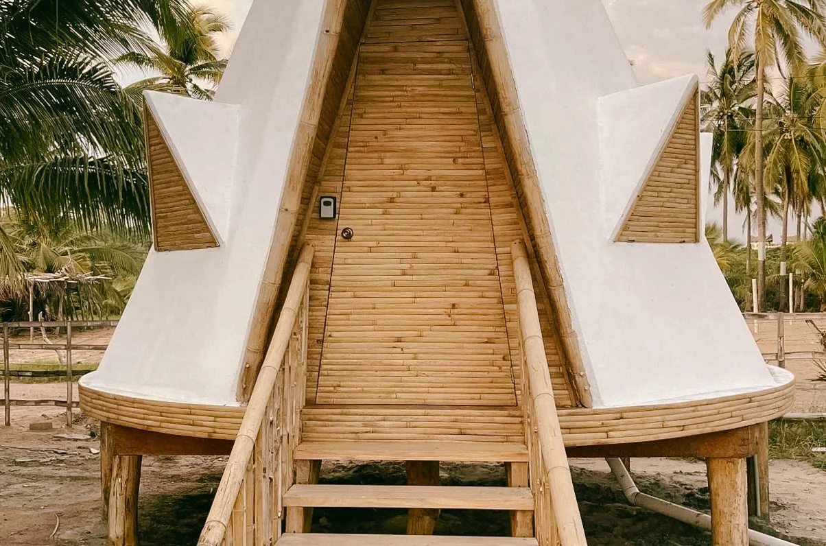 Beachfront housing; A small, modern teepee with a steep, pointed roof, built using bamboo and concrete, located outdoors with palm trees in the background.
