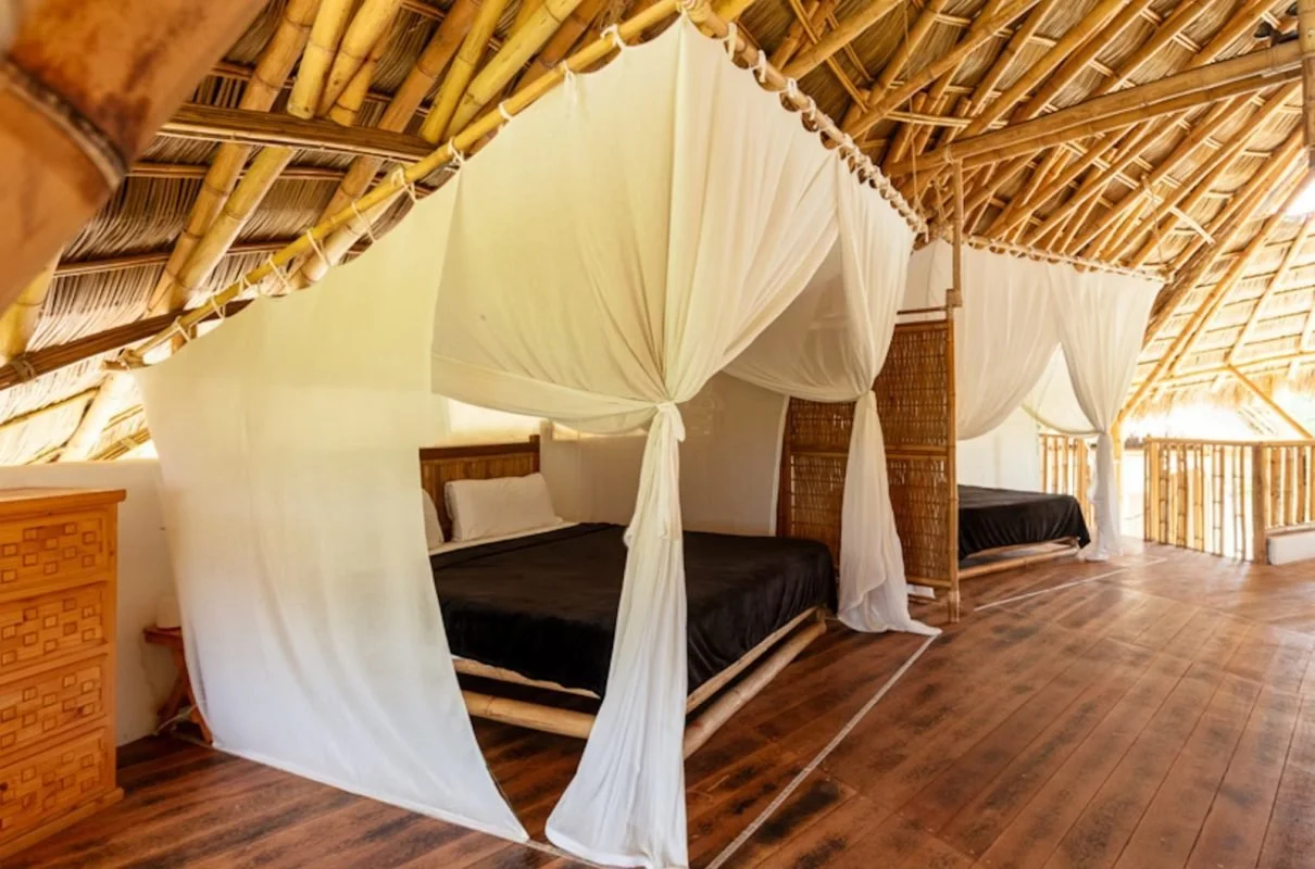 Villa Tortuga interior; Two beds in a bamboo hut with white canopy curtains, black bedding, and wooden flooring.
