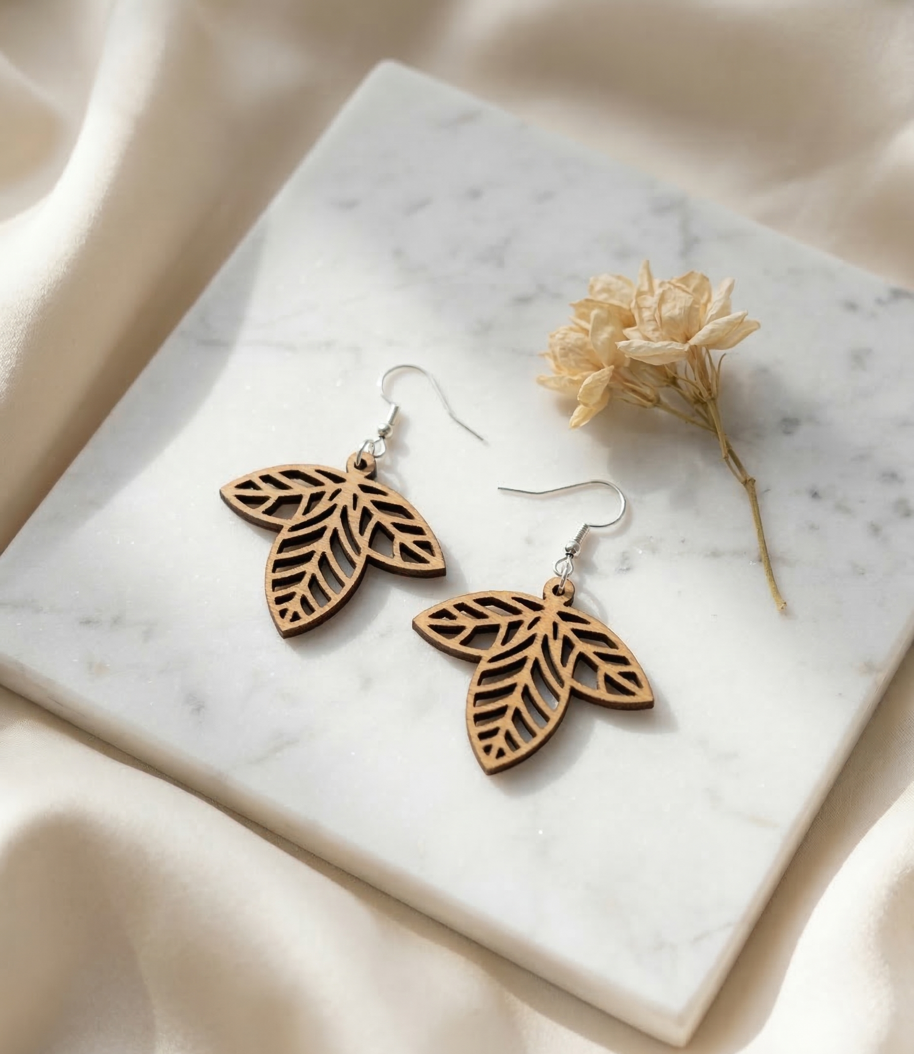 Wooden leaf-shaped earrings with intricate cutout design placed on a white marble surface, accompanied by a dried flower and a beige fabric background.