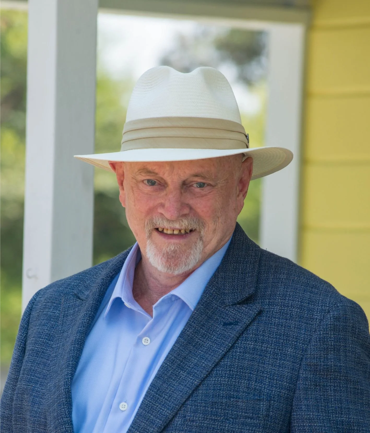 An elderly man wearing a white wide-brimmed hat, a light blue dress shirt, and a blue blazer, smiling outdoors near a yellow house with a porch.