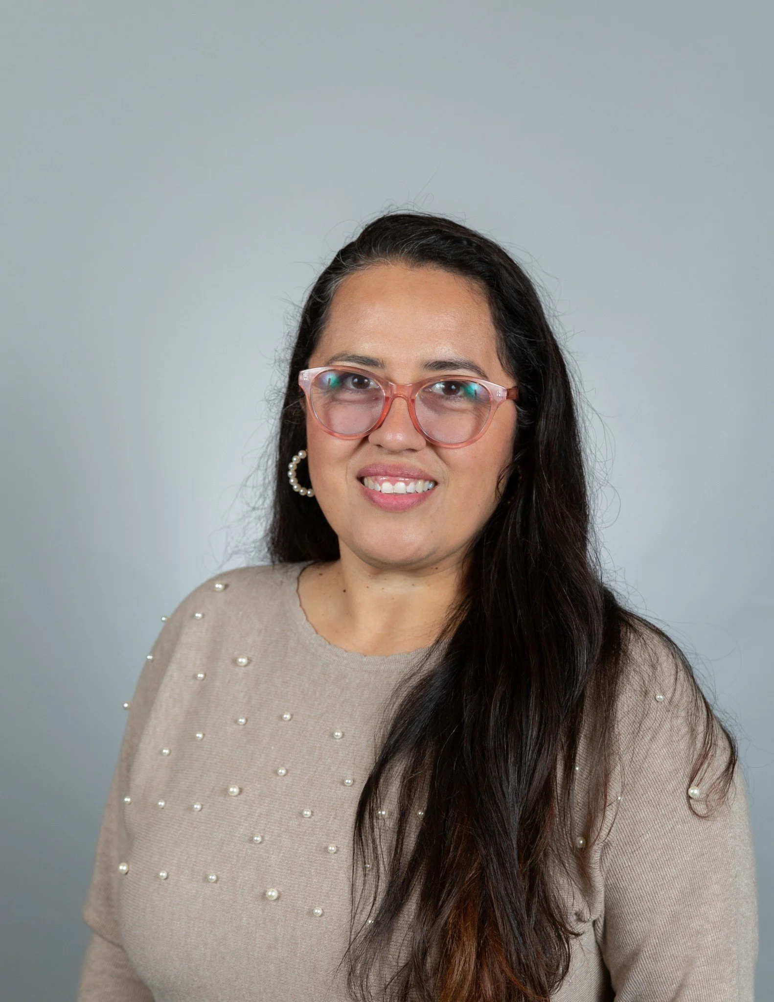 A woman with long dark hair wearing pink glasses, a beige sweater with pearl embellishments, and a pearl earring, smiling at the camera against a plain gray background.