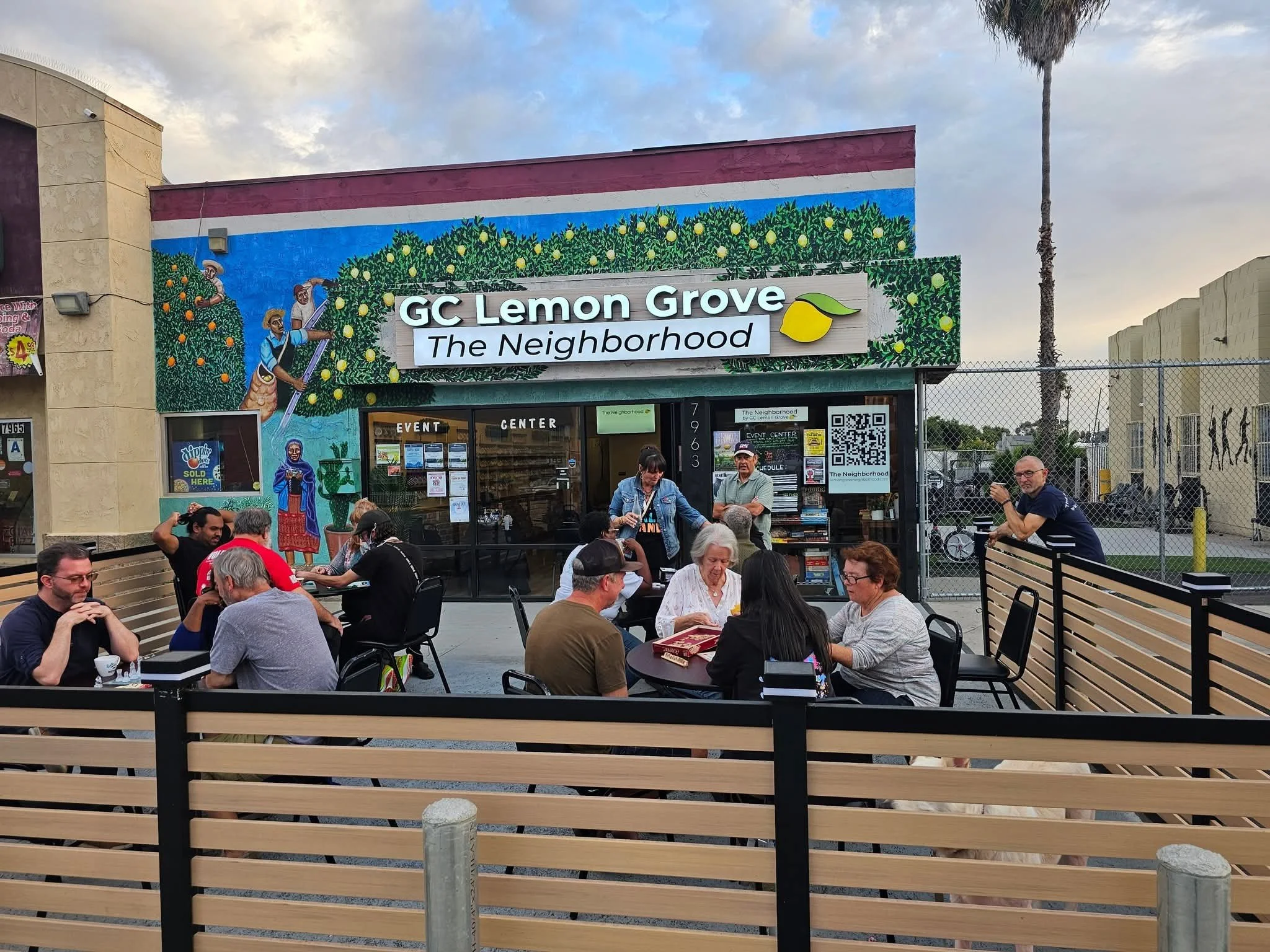 People sitting and standing outside "The Neighborhood" event center with a large, colorful mural on the building behind them. The mural depicts orange and lemon trees, with some figures picking lemon. The sign reads "GC Lemon Grove The Neighborhood."