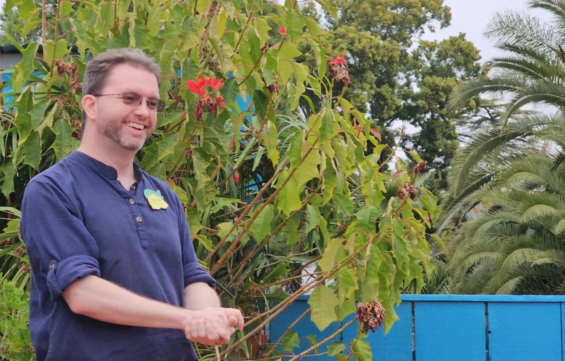 A smiling man with glasses and a beard wearing a navy blue shirt stands outdoors in a garden. There are green trees, plants, and a blue fence in the background.