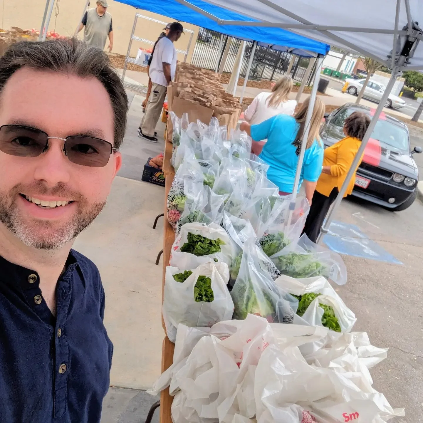 A man taking a selfie at the Lemon Grove Food Distribution with a table of fresh vegetables in plastic bags. There are several people in the background working under a blue canopy, with cars parked nearby.