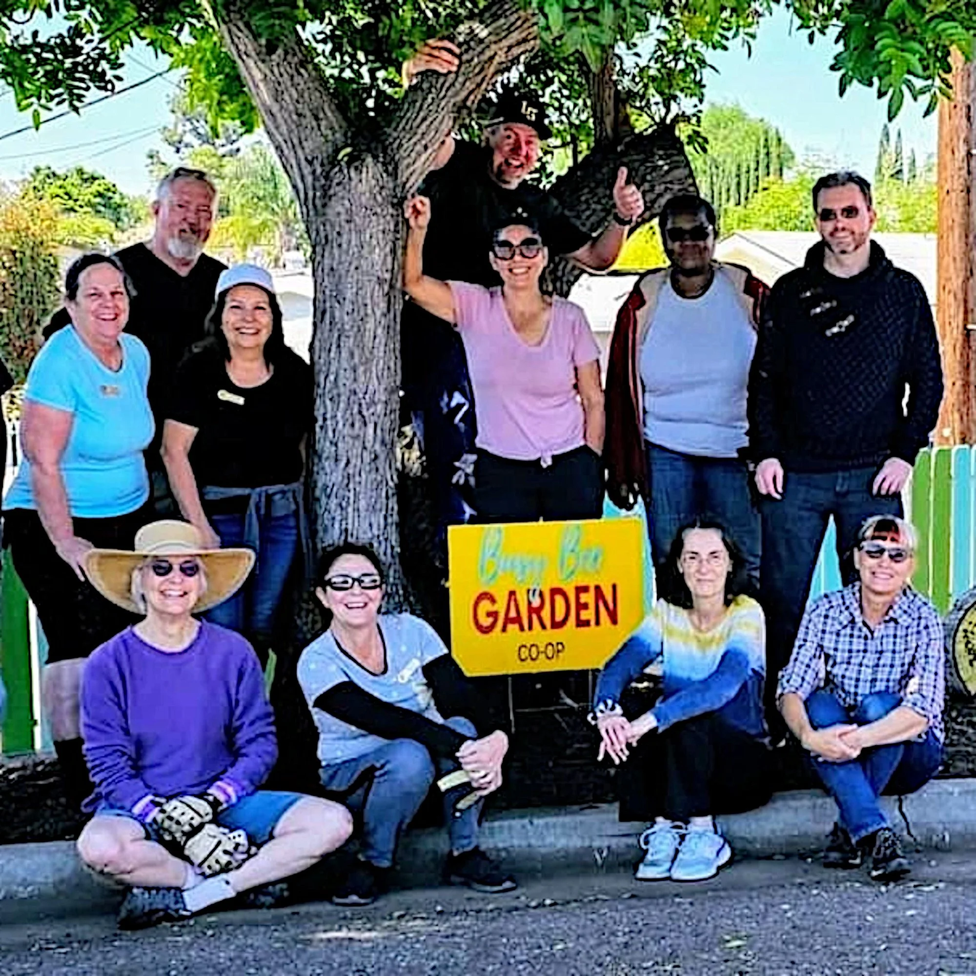Group of people celebrating at a community gardening event, some wearing sunglasses and smiling, with a colorful sign that reads 'BUSY BEE GARDEN CO-OP'.