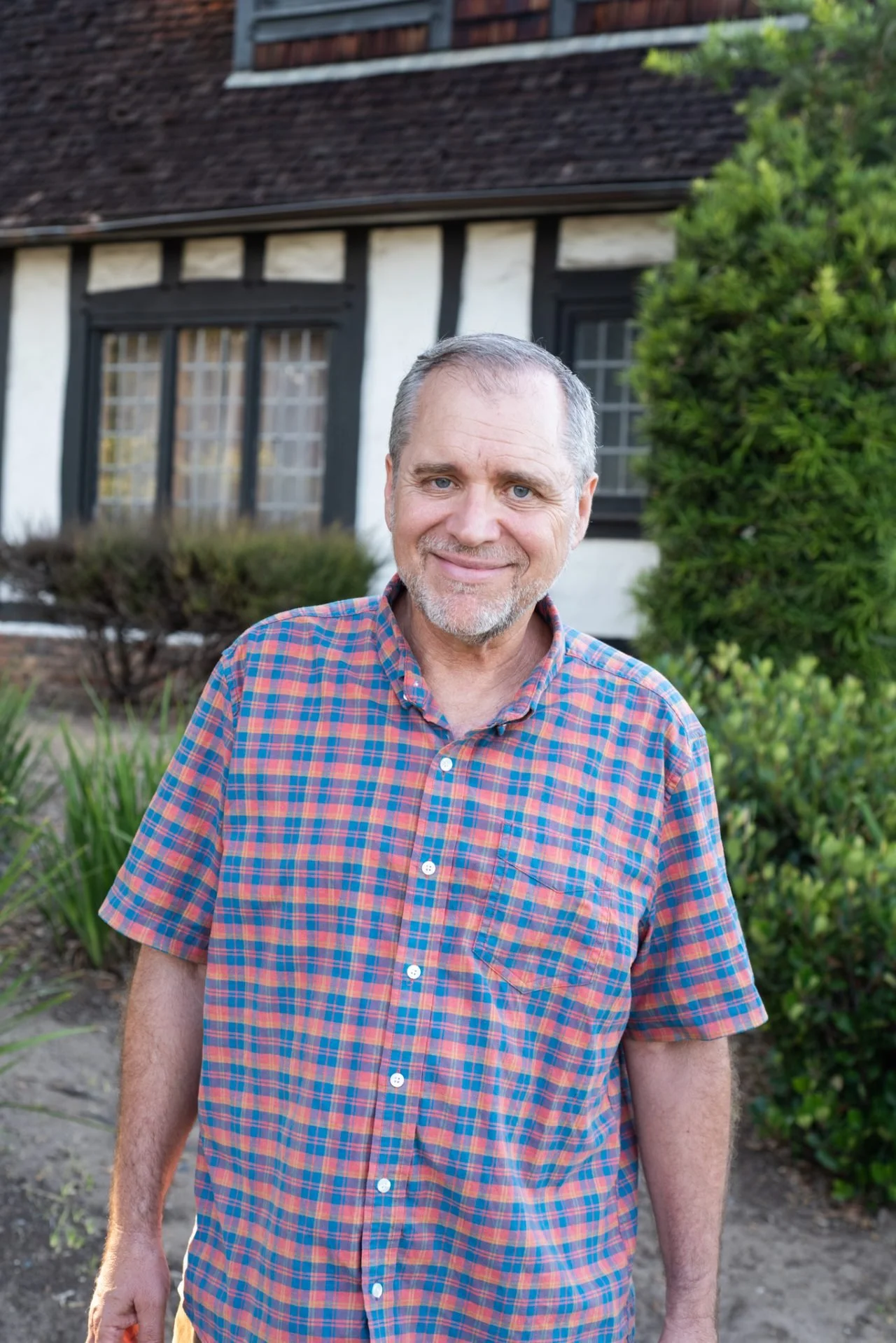 A middle-aged man with gray hair and a beard smiling outdoors in front of a house with black and white timber framing, surrounded by greenery.