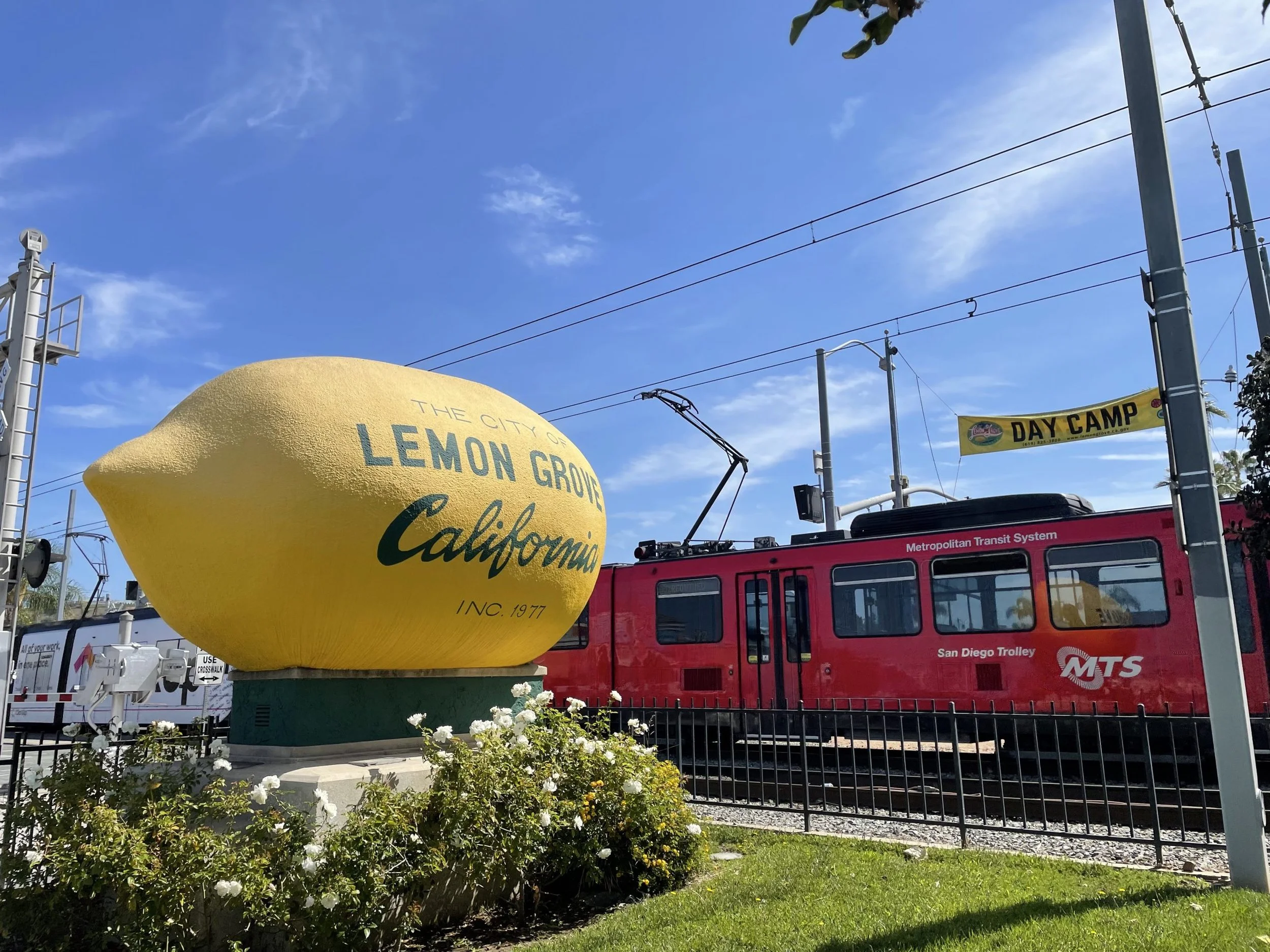 A large lemon-shaped sign reads 'The City of Lemon Grove California, Inc. 1977.' Next to it is a red San Diego Trolley train car on tracks, with a yellow banner overhead that says 'Day Camp.' There are some flowers in the foreground and blue sky above.