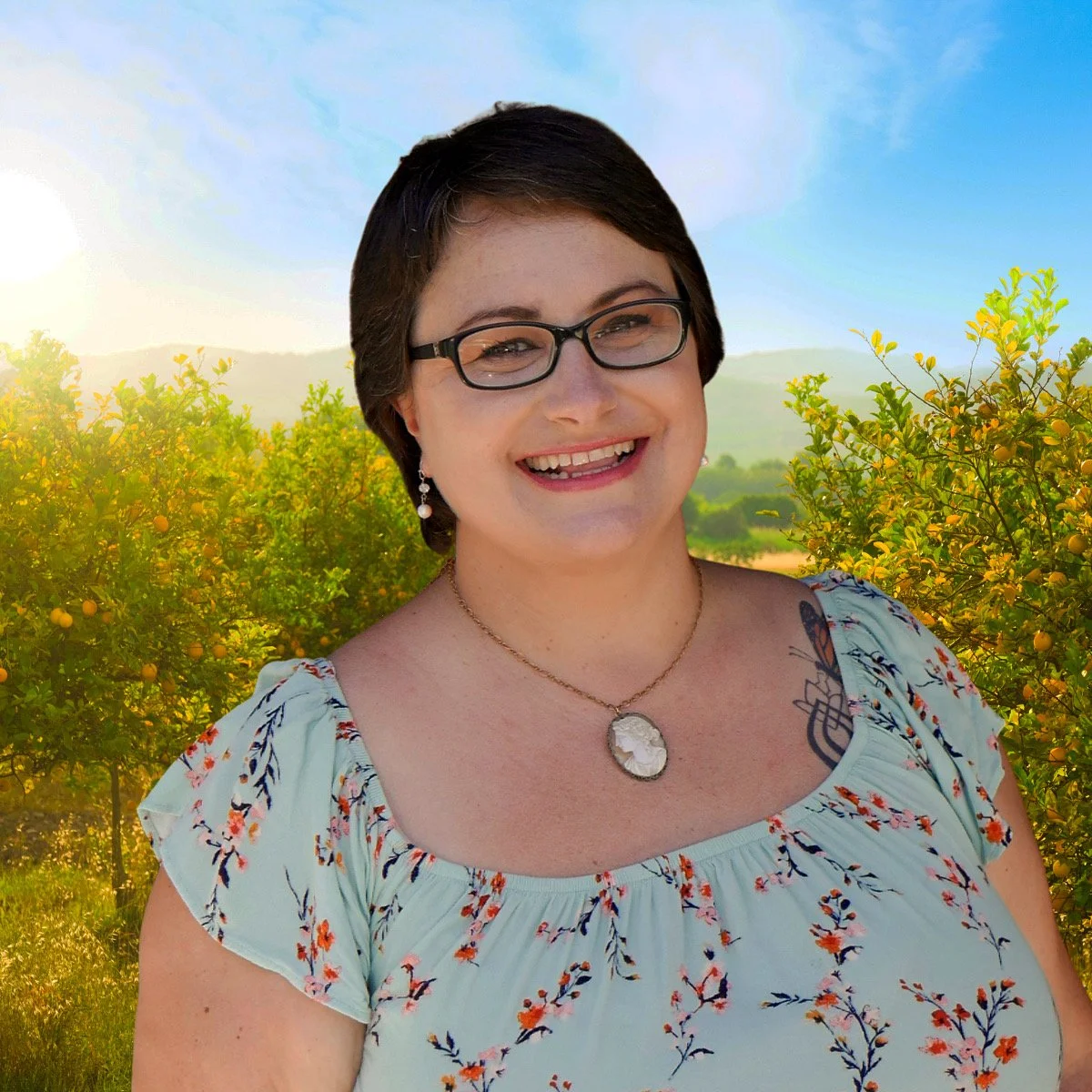 A woman with short dark hair and glasses smiling outdoors, with a backdrop of lemon trees and a blue sky.