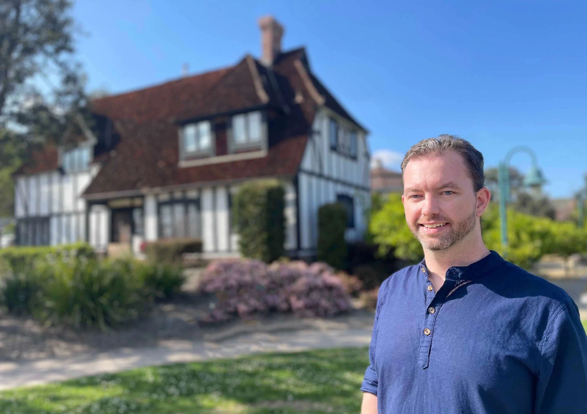 A man with short brown hair and a beard, smiling, standing outdoors in front of a blurred house and garden on a bright sunny day.