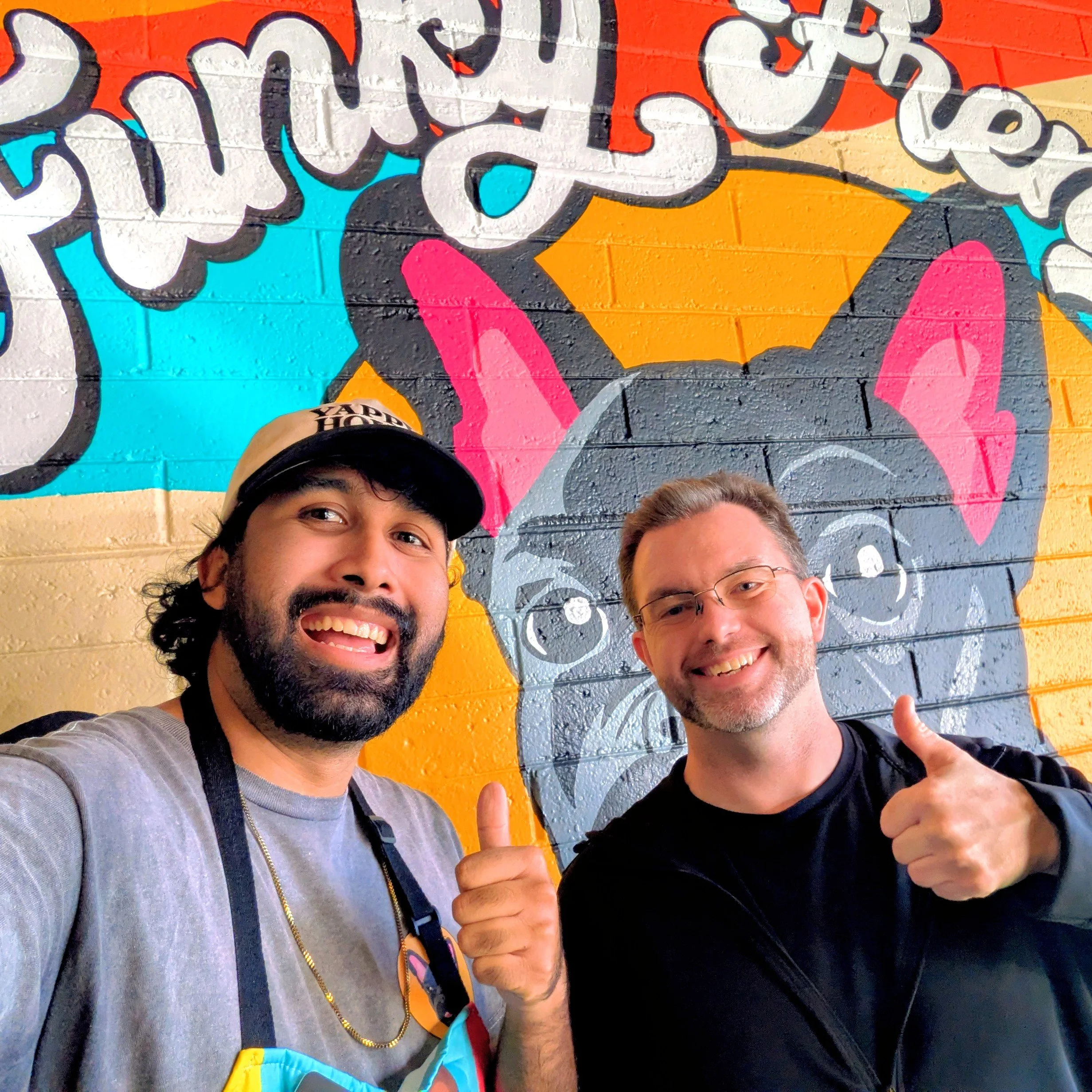 Two men smiling and giving thumbs up in front of a colorful mural of a French Bulldog on a brick wall.