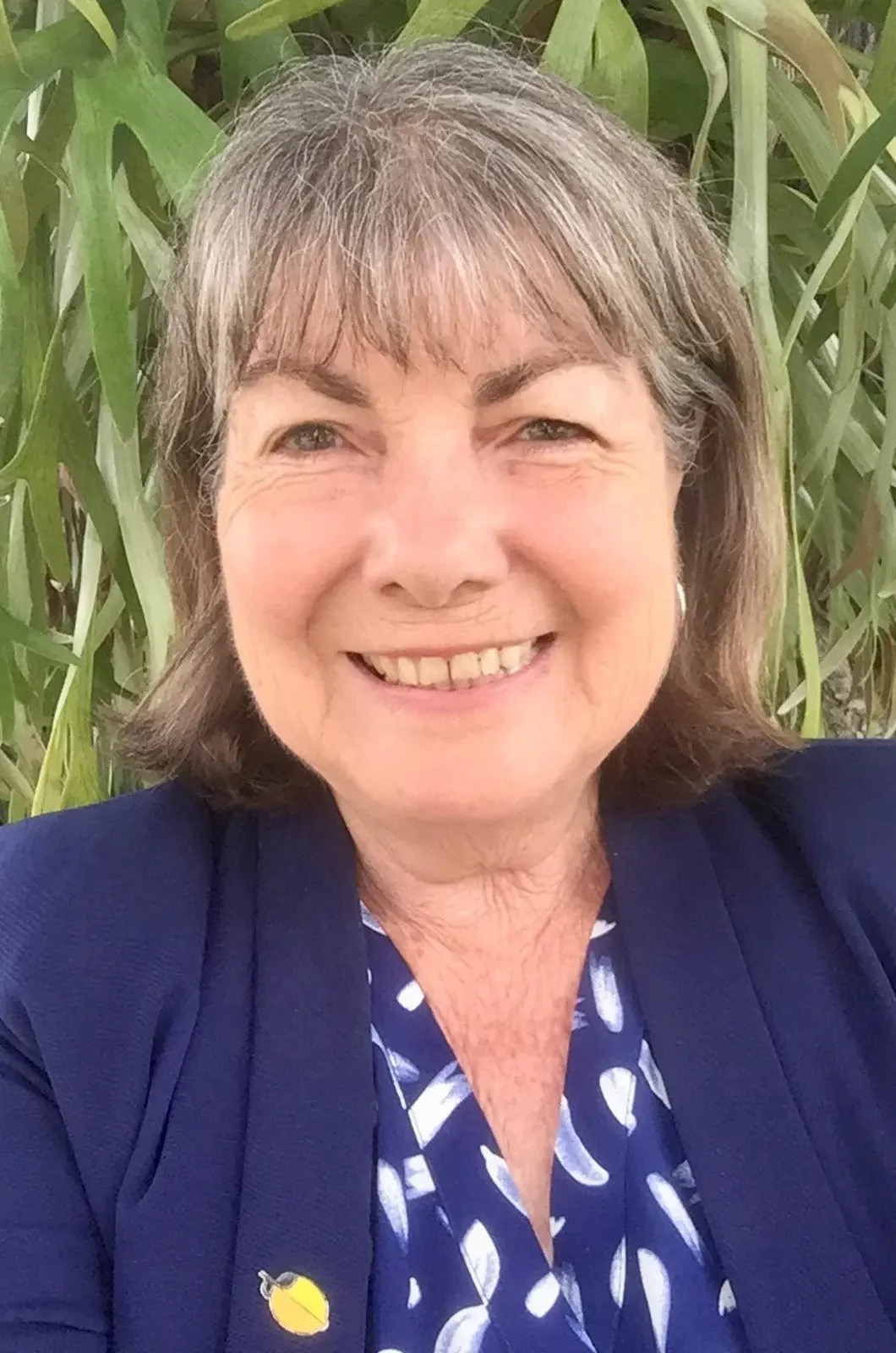 A smiling woman with short gray hair wearing a dark blue blazer and a patterned shirt, against a background of green plants.