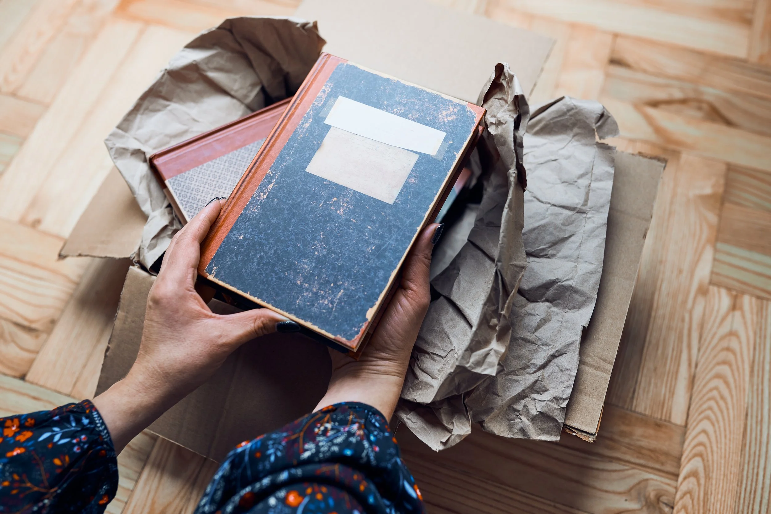 A person wearing a dark floral-patterned blouse is holding a vintage-style blue and orange notebook with blank white labels on the cover, over crumpled brown paper inside a cardboard box on a wooden surface.