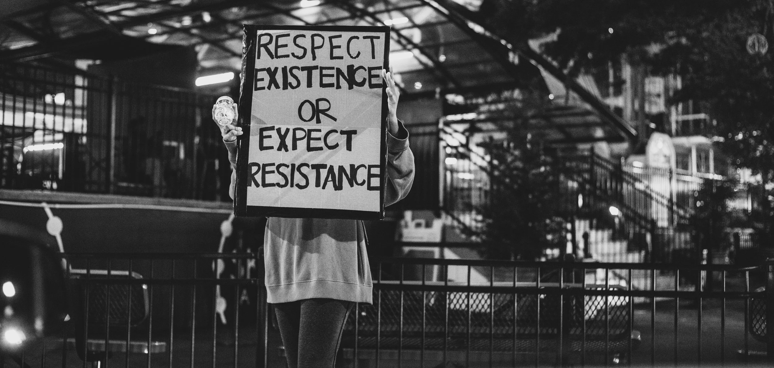 A person holding a sign that reads 'Respect Existence or Expect Resistance' at night in an outdoor urban setting with a metal fence and city buildings in the background.