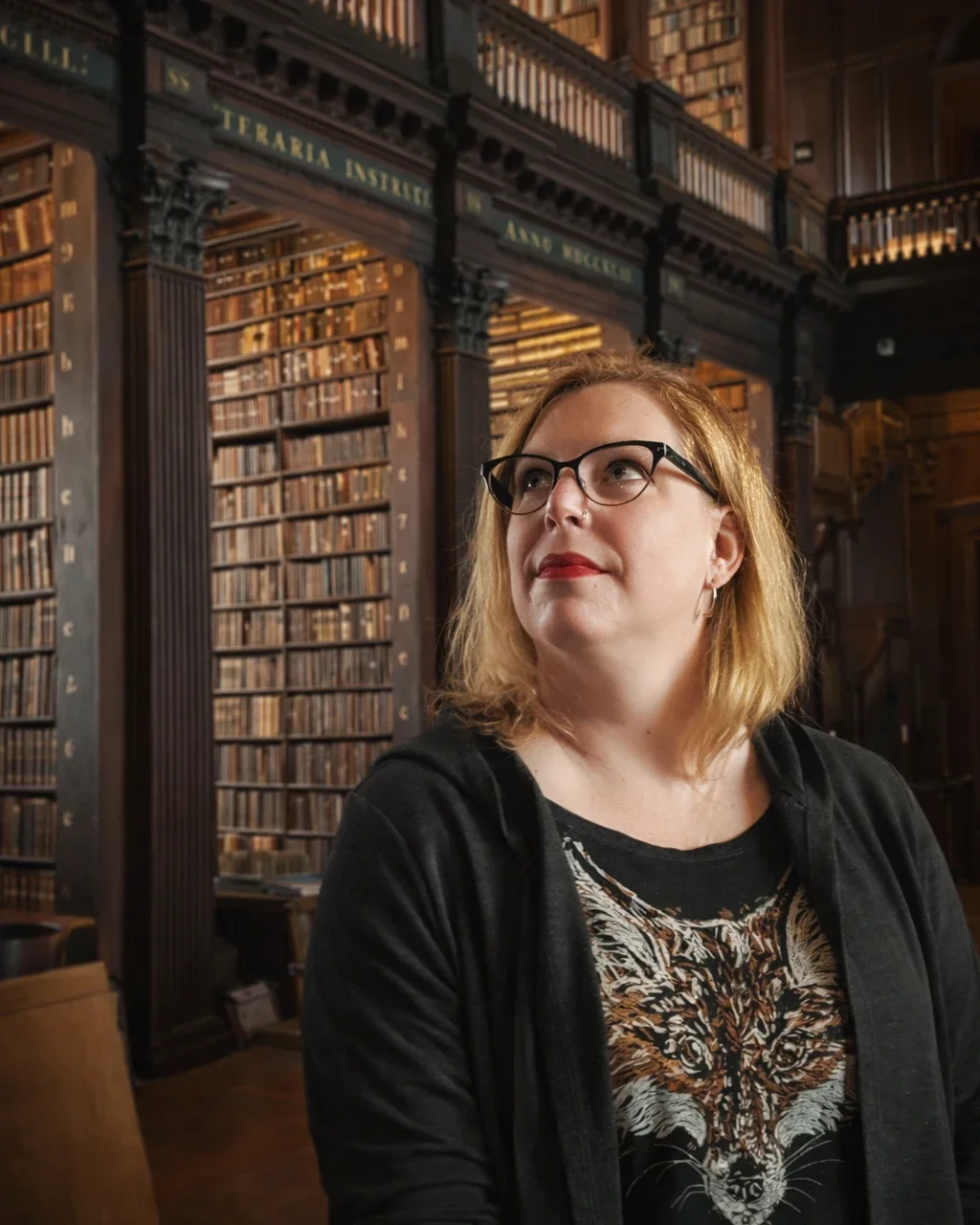 A woman with glasses and red hair wearing a black jacket and a tiger print shirt, standing inside a grand old library with tall wooden bookshelves filled with books.