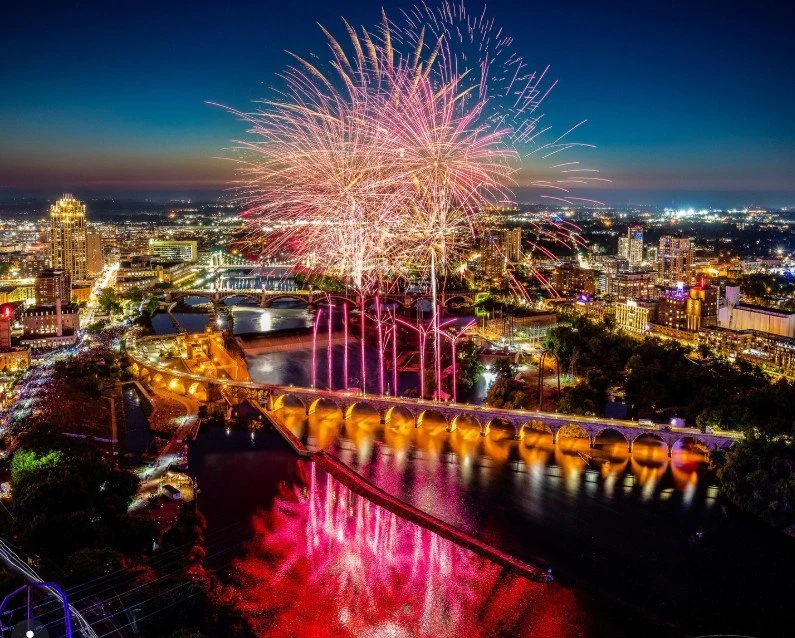 Colorful fireworks display over a city skyline at night, reflecting on a river with a bridge.