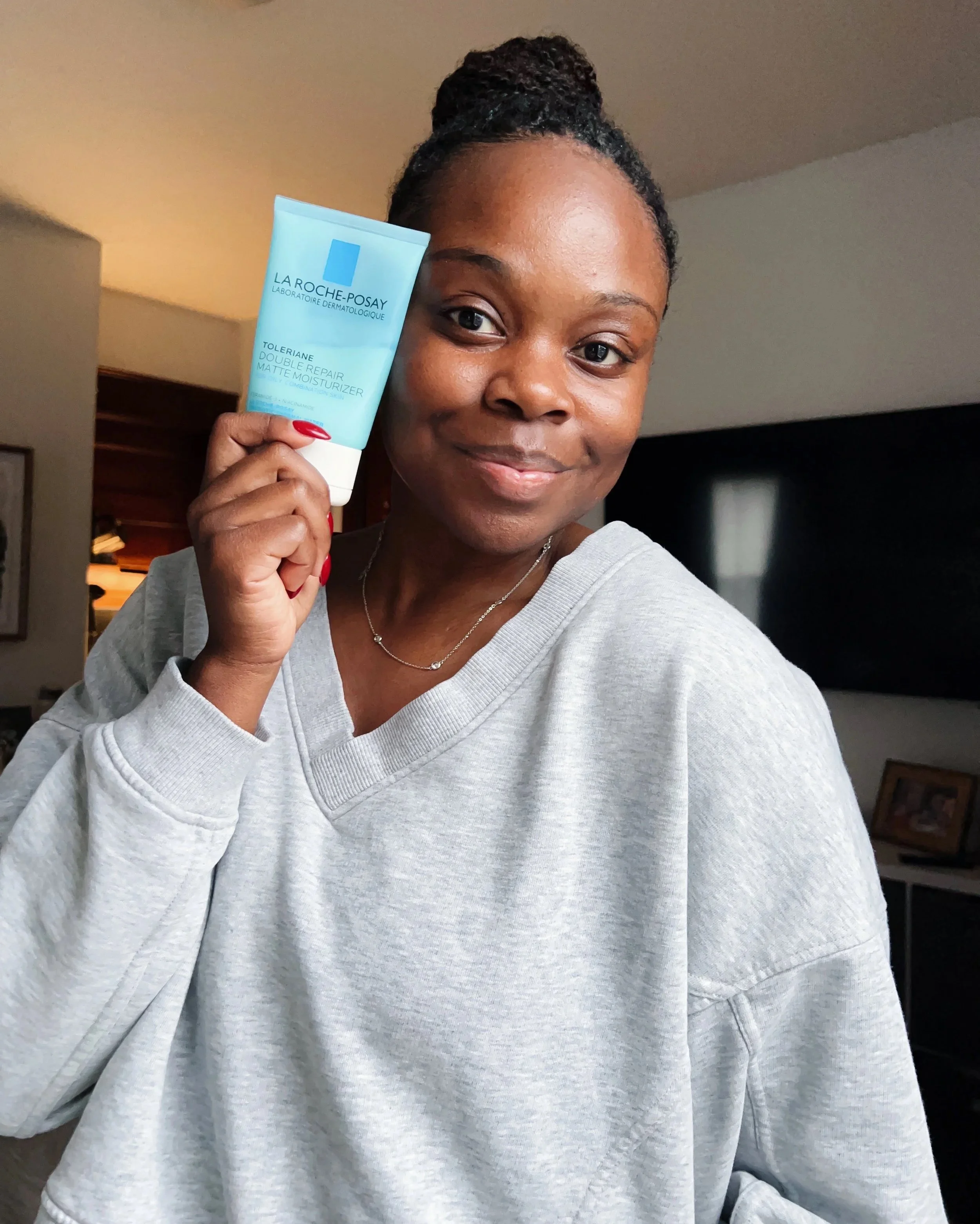 A woman with braided hair in a bun wearing a light gray sweatshirt, holding a blue tube of La Roche-Posay moisturizer, smiling.