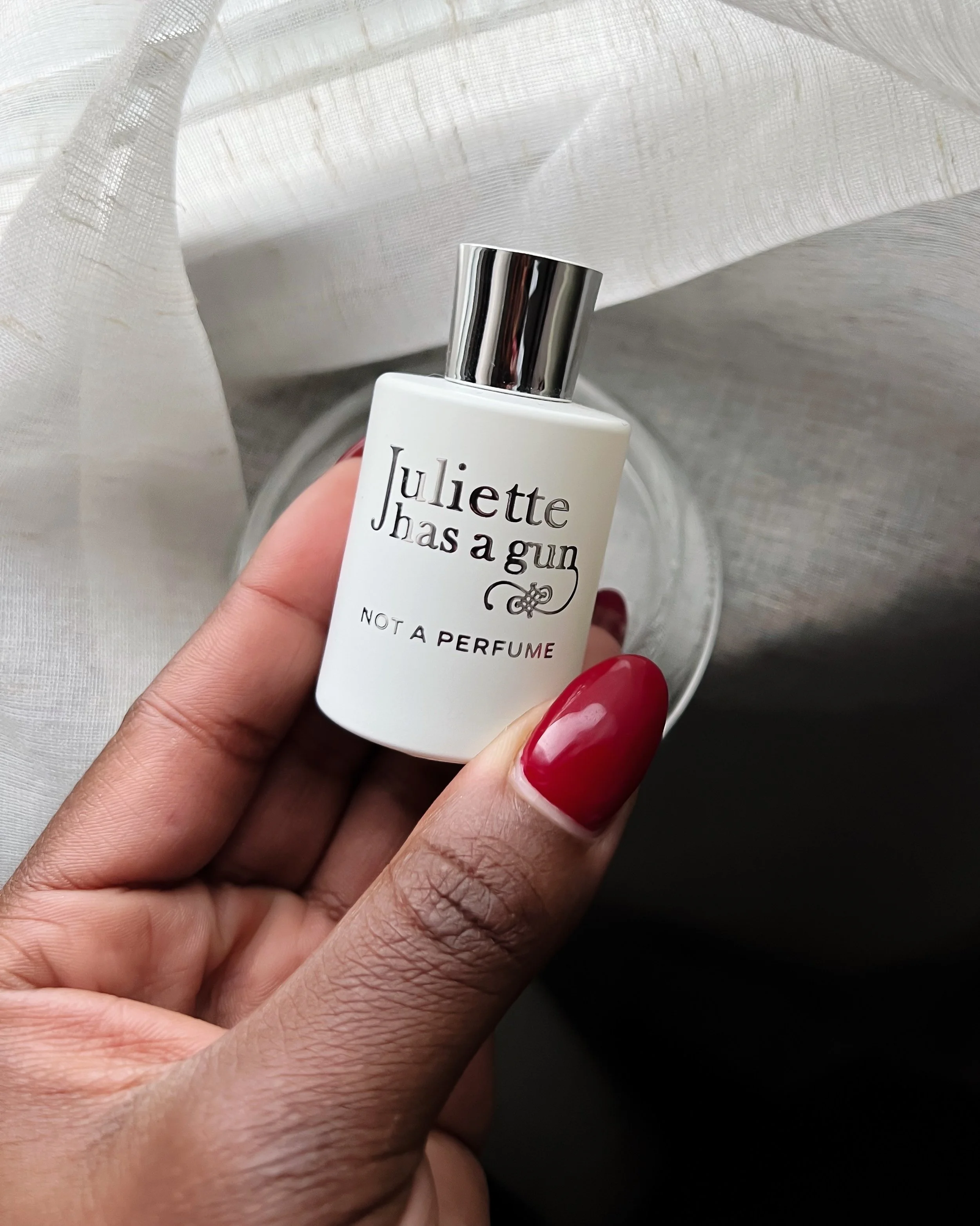 A person's hand with red nail polish holding a white perfume bottle that says 'Juliette Has a Gun Not a Perfume'.