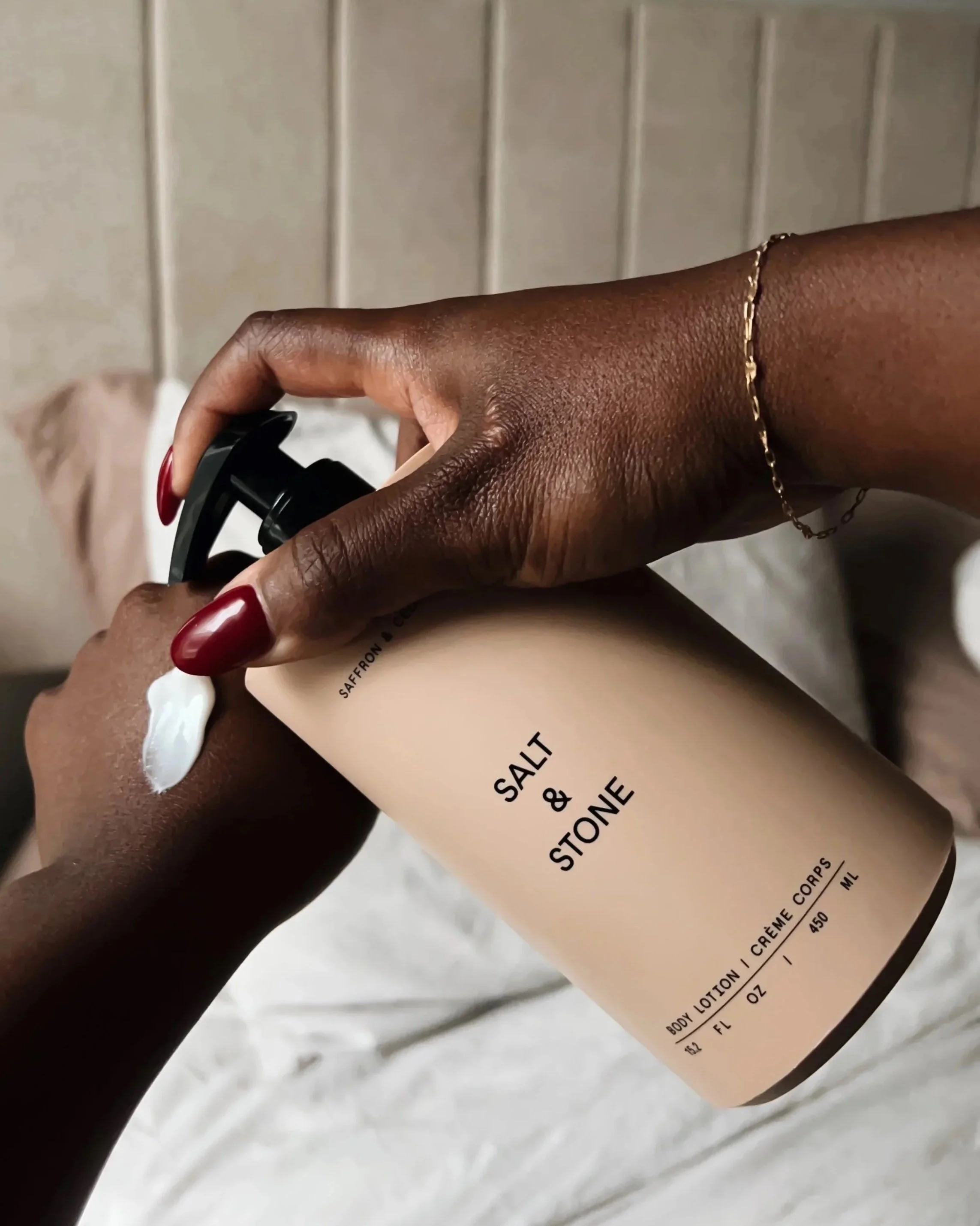 Person with dark skin applying white lotion from a beige bottle labeled 'Salt & Stone Body Lotion' on their arm.