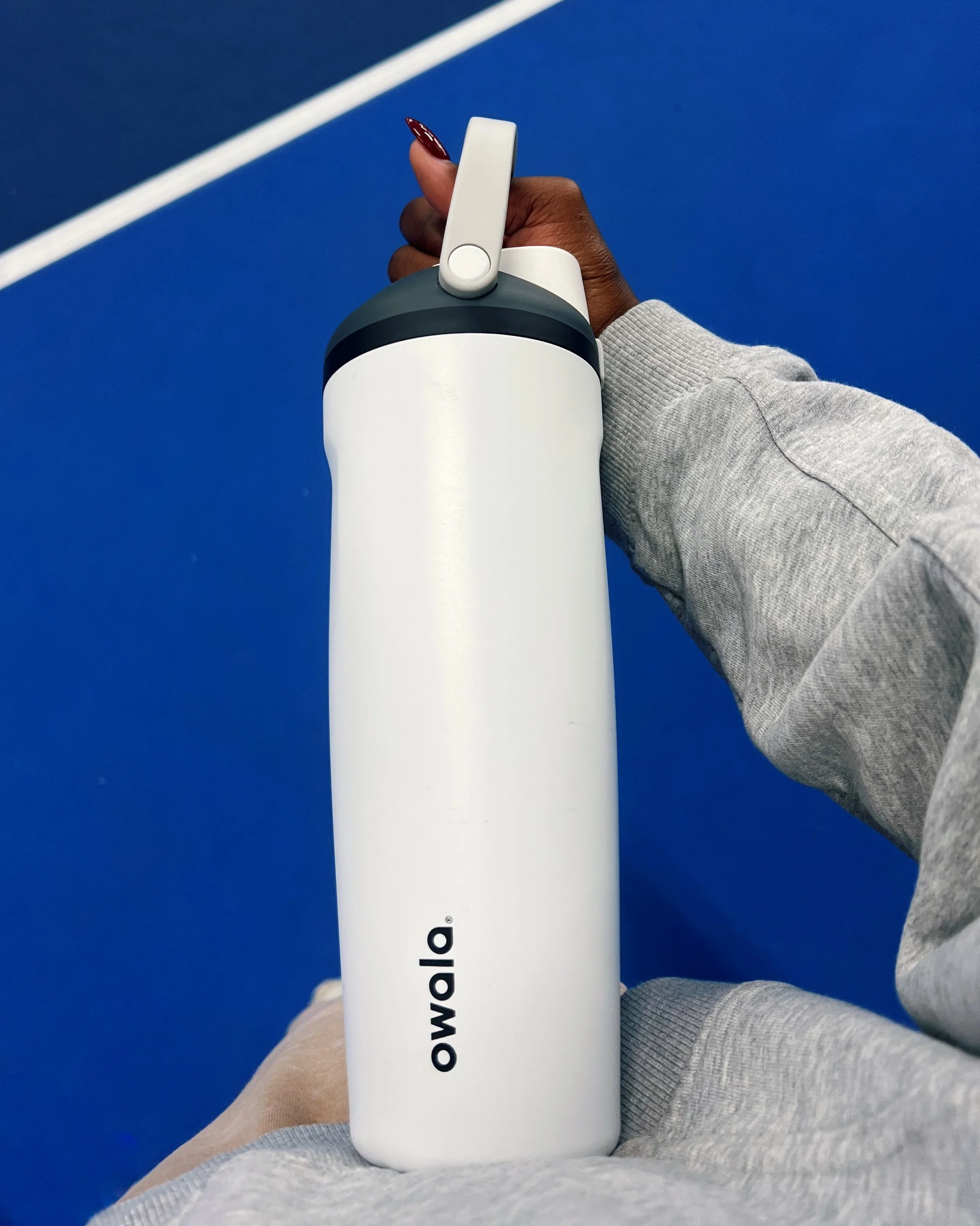 A person holding a white Owalla water bottle on a blue surface, wearing gray clothing with painted nails.