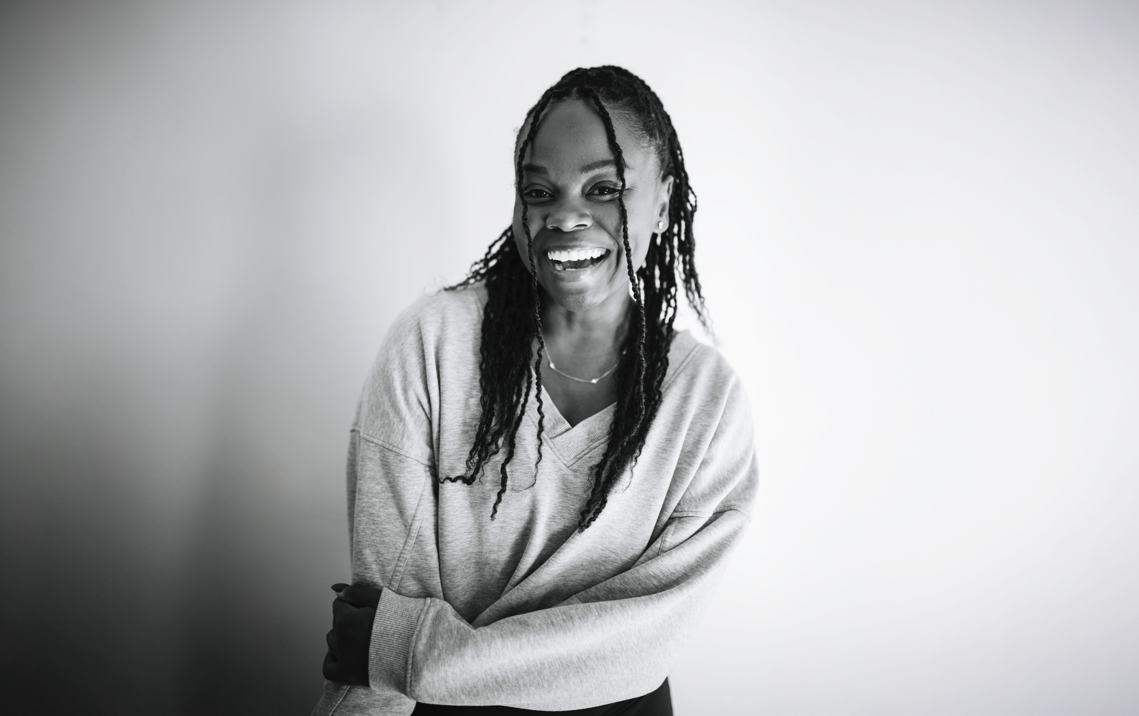 Black and white photo of a smiling woman with dreadlocks, wearing a casual sweatshirt and jewelry, standing against a plain background.