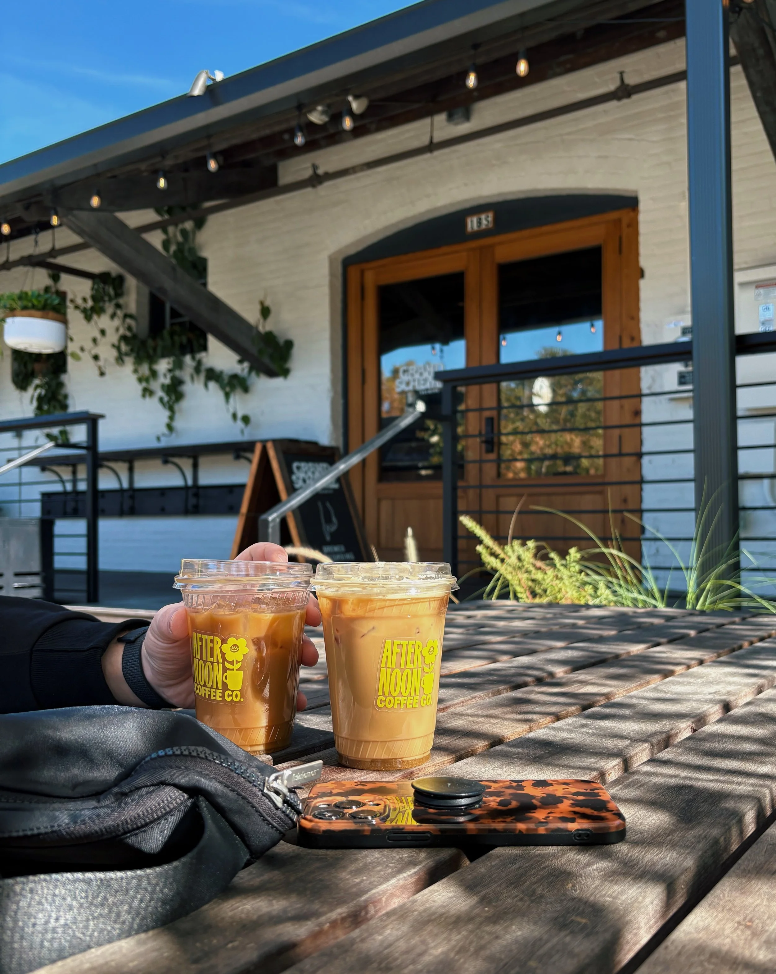 Two iced coffee drinks on a wooden table outside a coffee shop with a white brick exterior, wooden door, and potted plants.