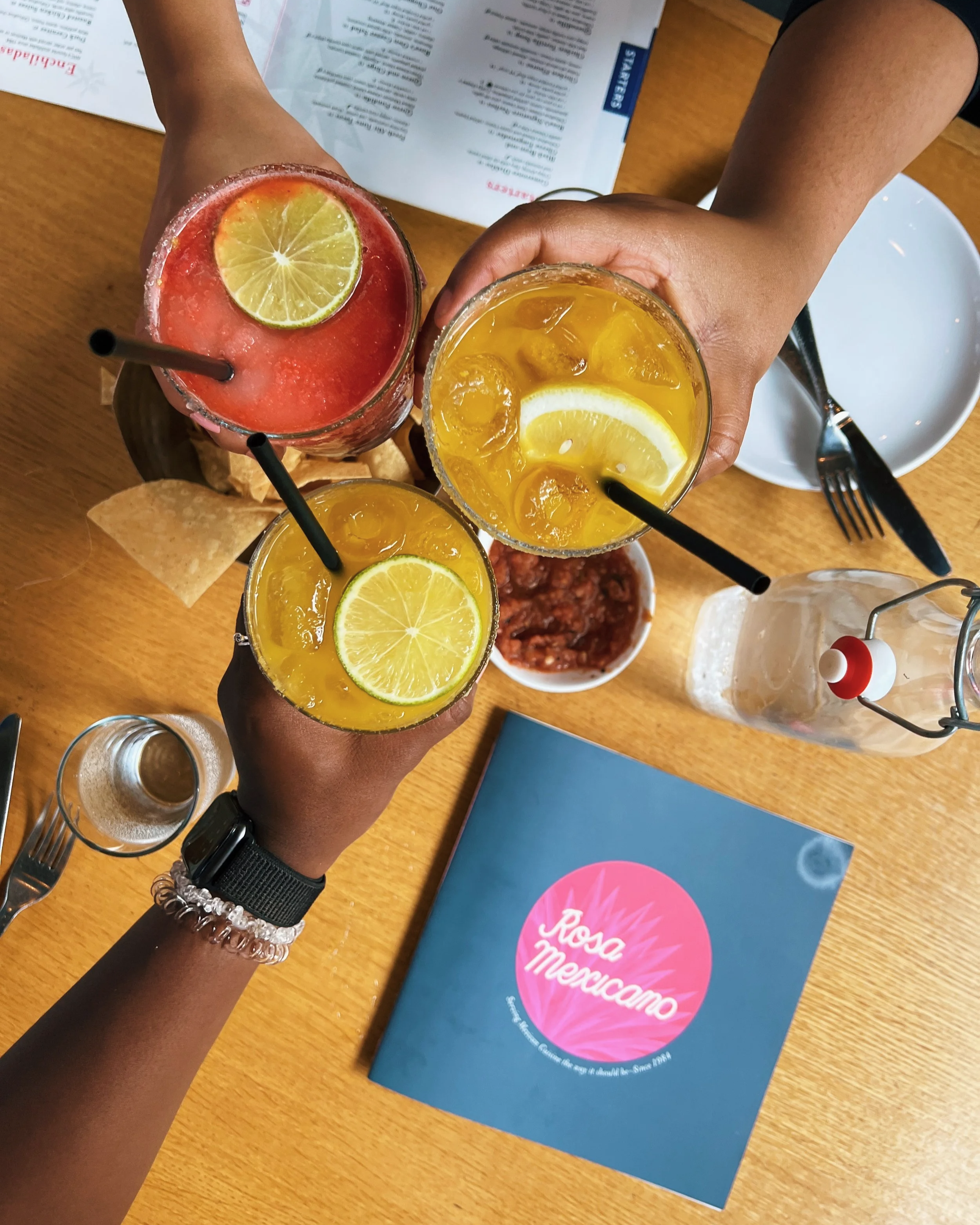 Three people holding glasses of colorful margaritas with lime slices, toasting over a wooden table with a blue menu that says 'Rosa Mexicano'.